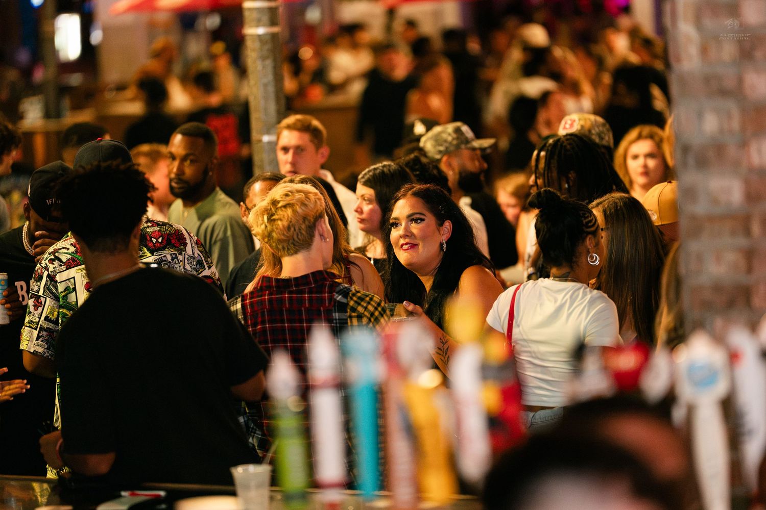 Smiling woman in plaid shirt mingling in a lively outdoor bar crowd.