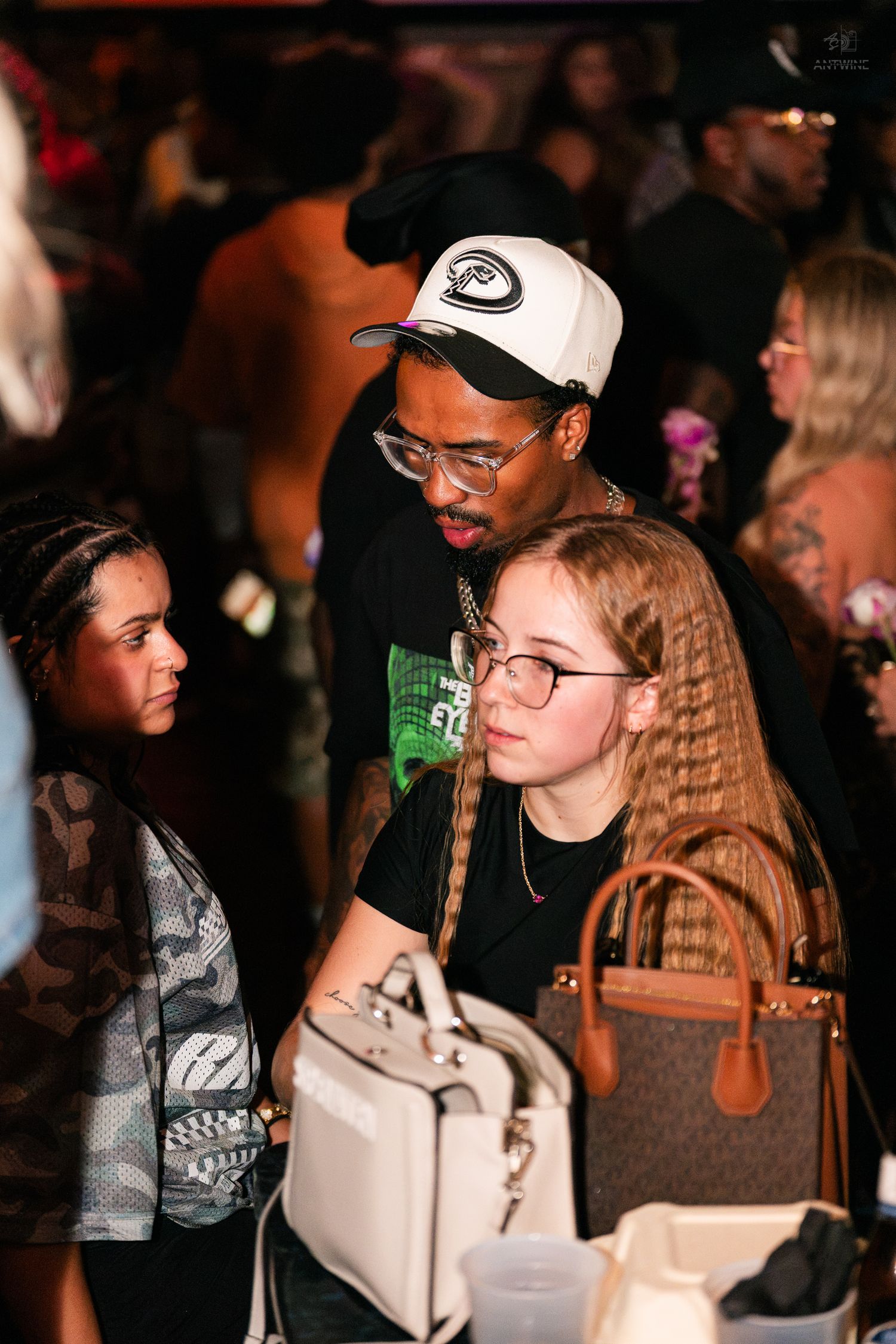 Man in white cap and glasses leans toward young woman with braids at a crowded nightclub event.