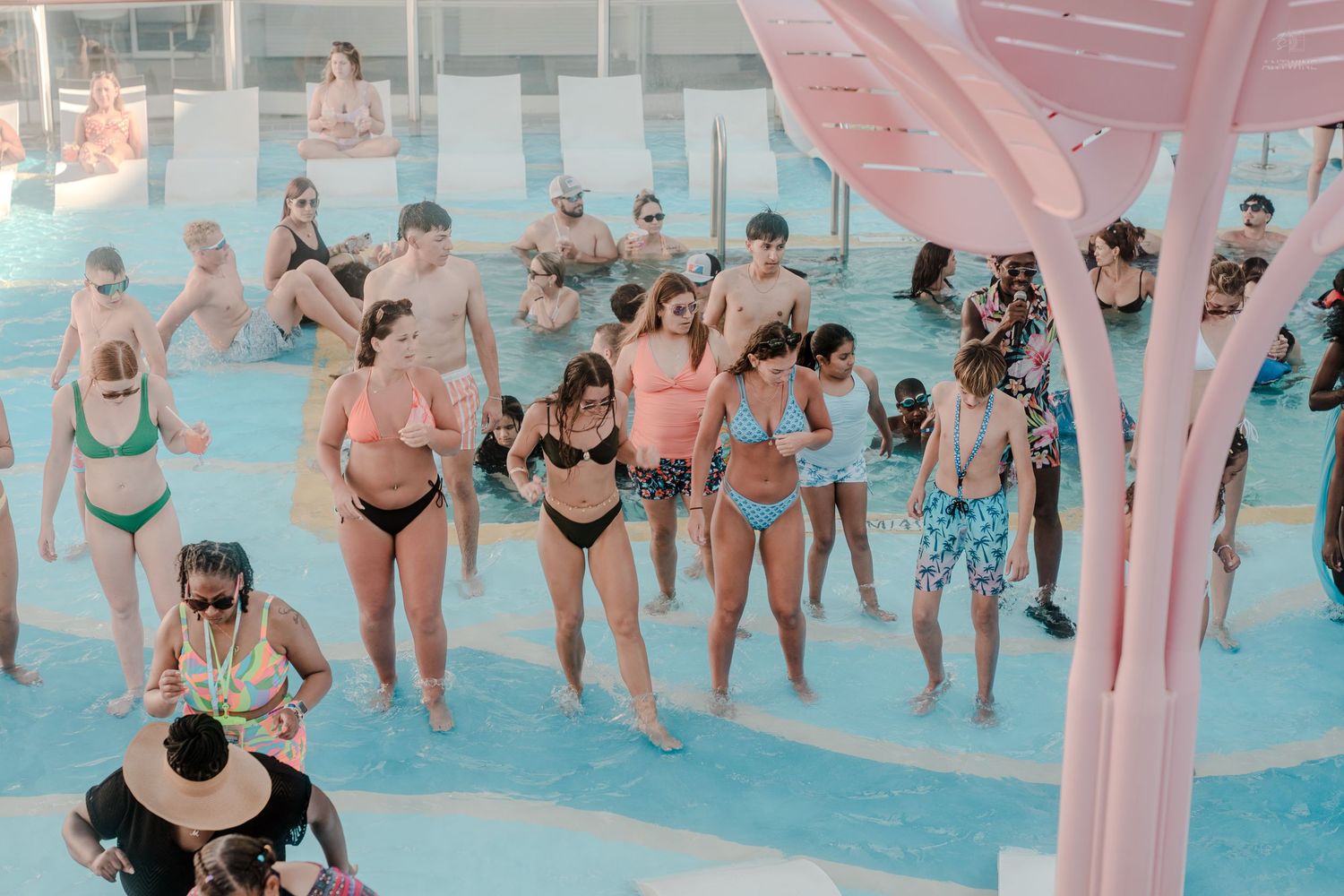 Group of cruise ship guests in swimwear dancing poolside near a pink decorative tree structure.