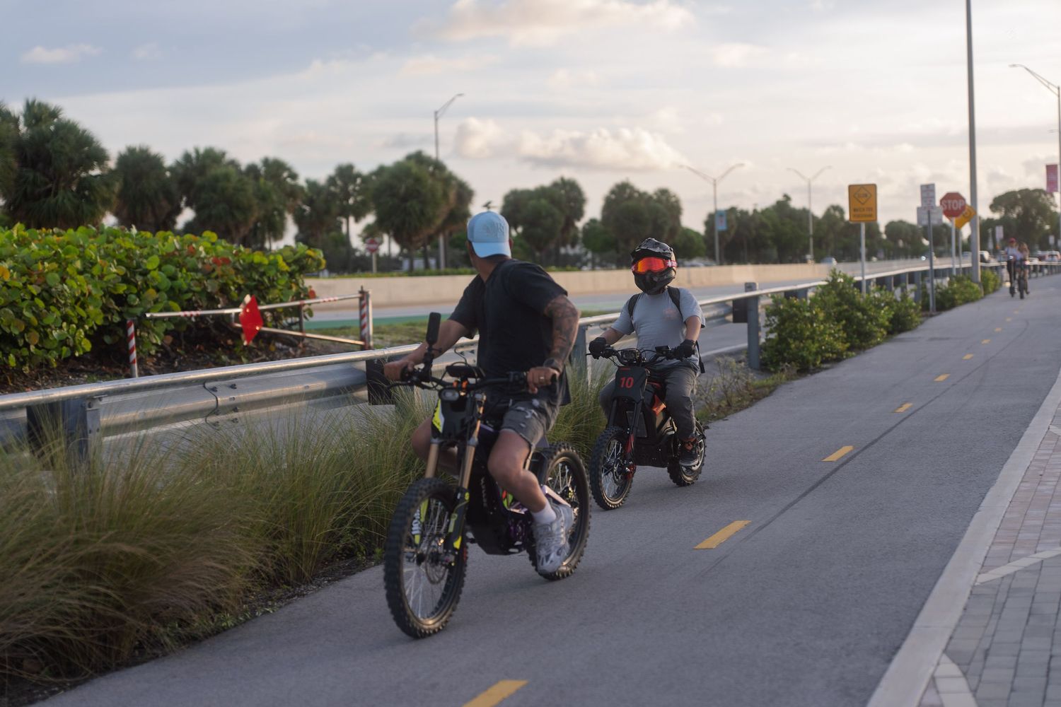 Two cyclists riding electric bikes along a scenic waterfront bike path lined with tropical palm trees at sunset.