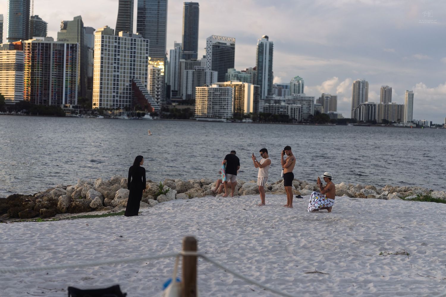Group of people gathered on a small sandy beach peninsula with Miami city skyline in the background.