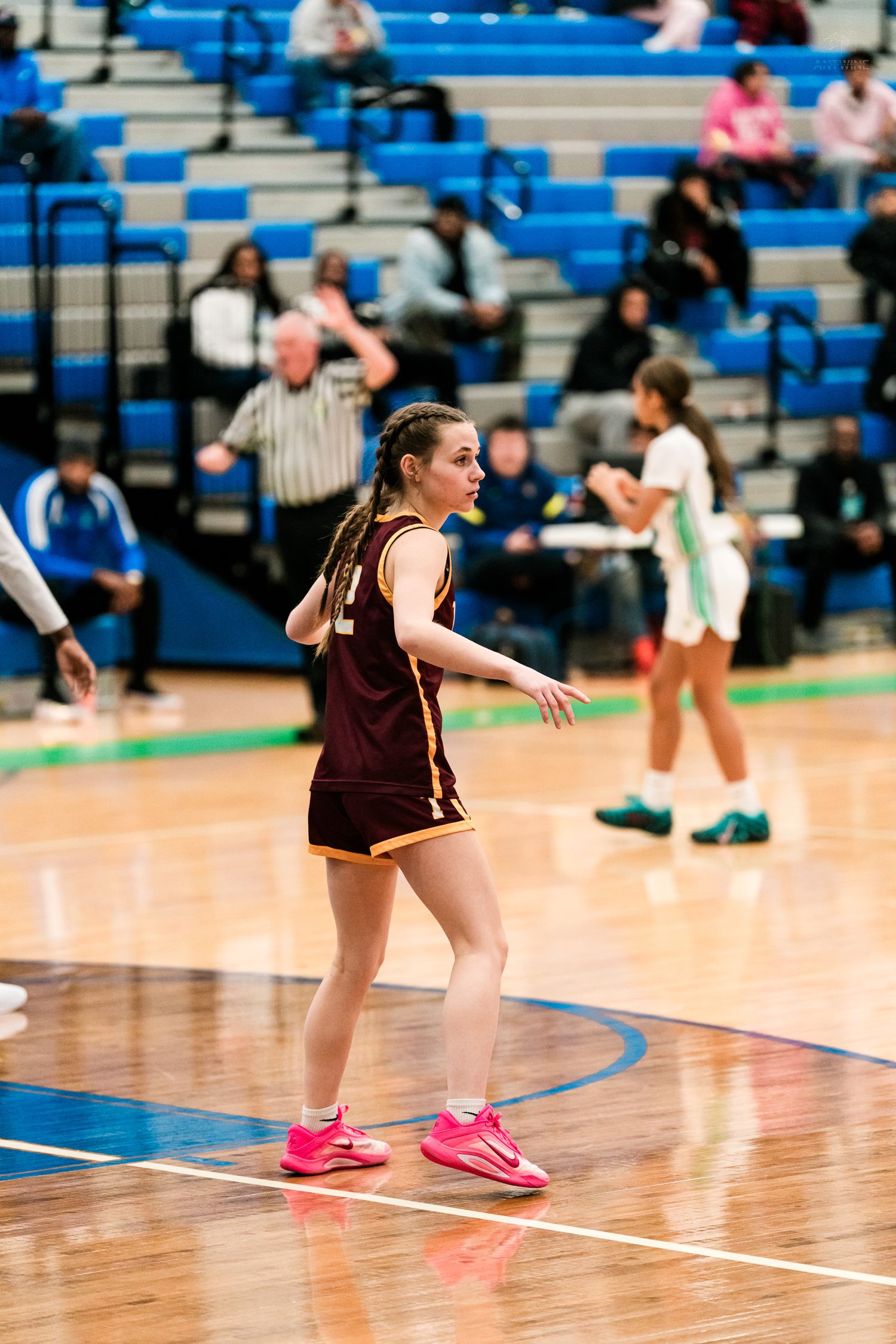 Female basketball player in maroon uniform with pink sneakers stands ready on a polished indoor court during a high school game.