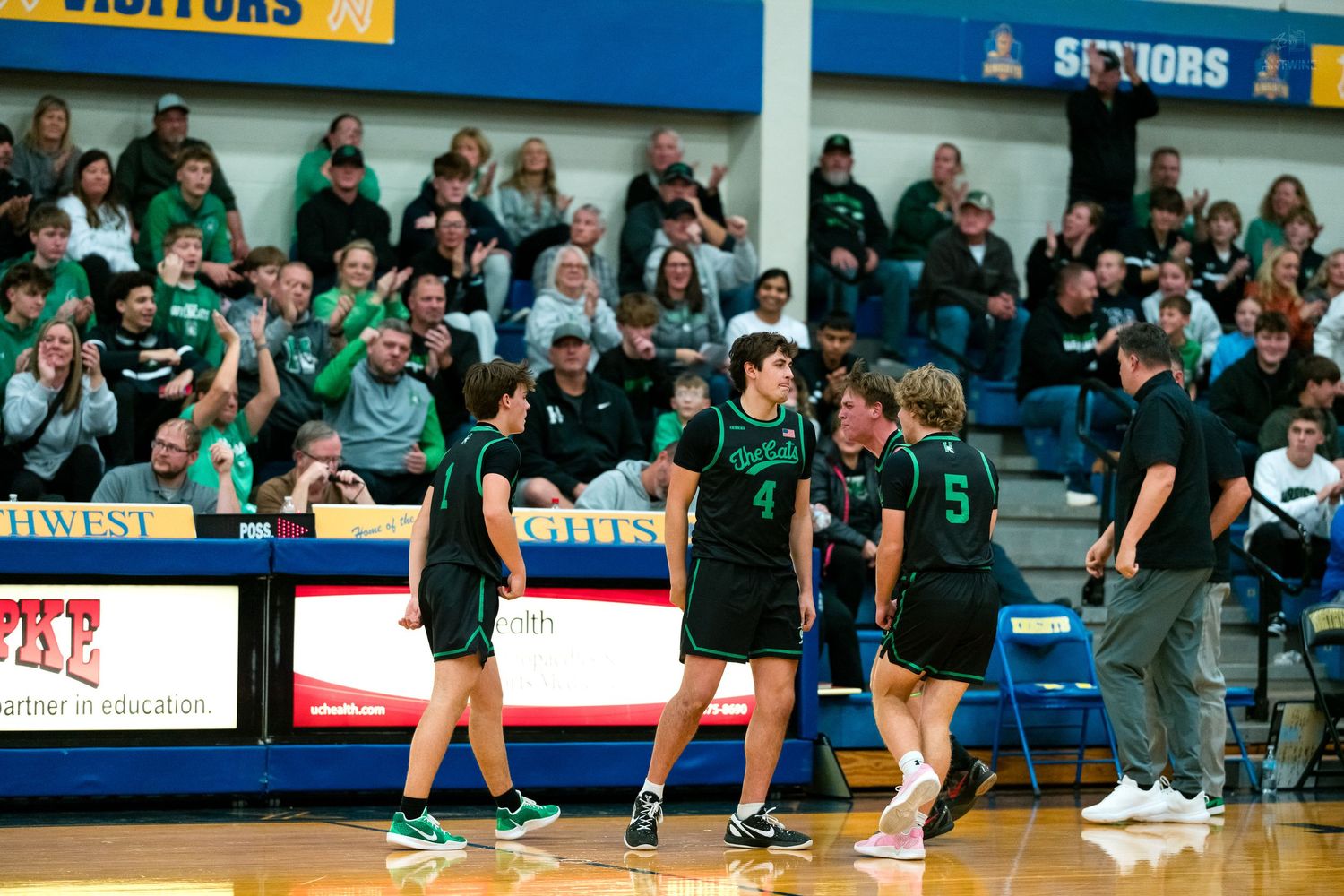 Three green uniformed basketball players celebrate on court with fans cheering in background.