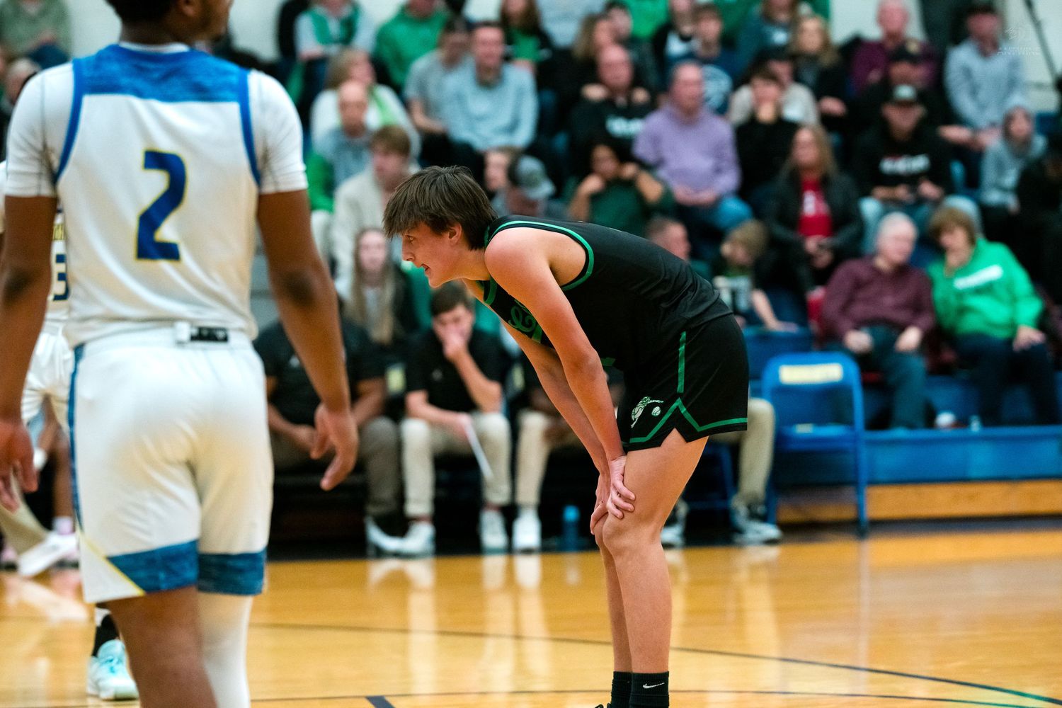 High school basketball player in green and black uniform bends forward during a break in play on the court.