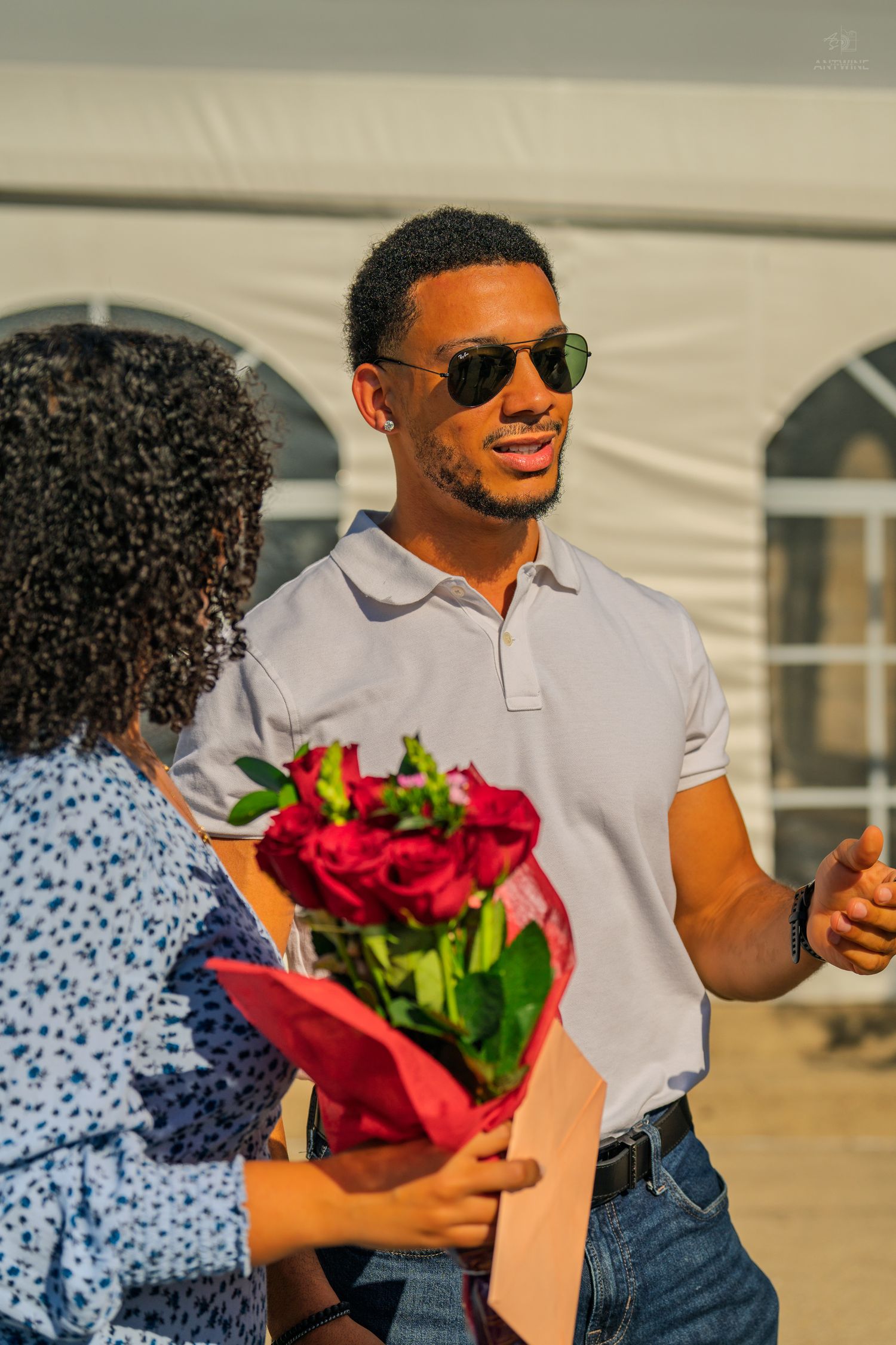 A man in sunglasses and white polo gestures while talking to a woman holding a bouquet of red roses.