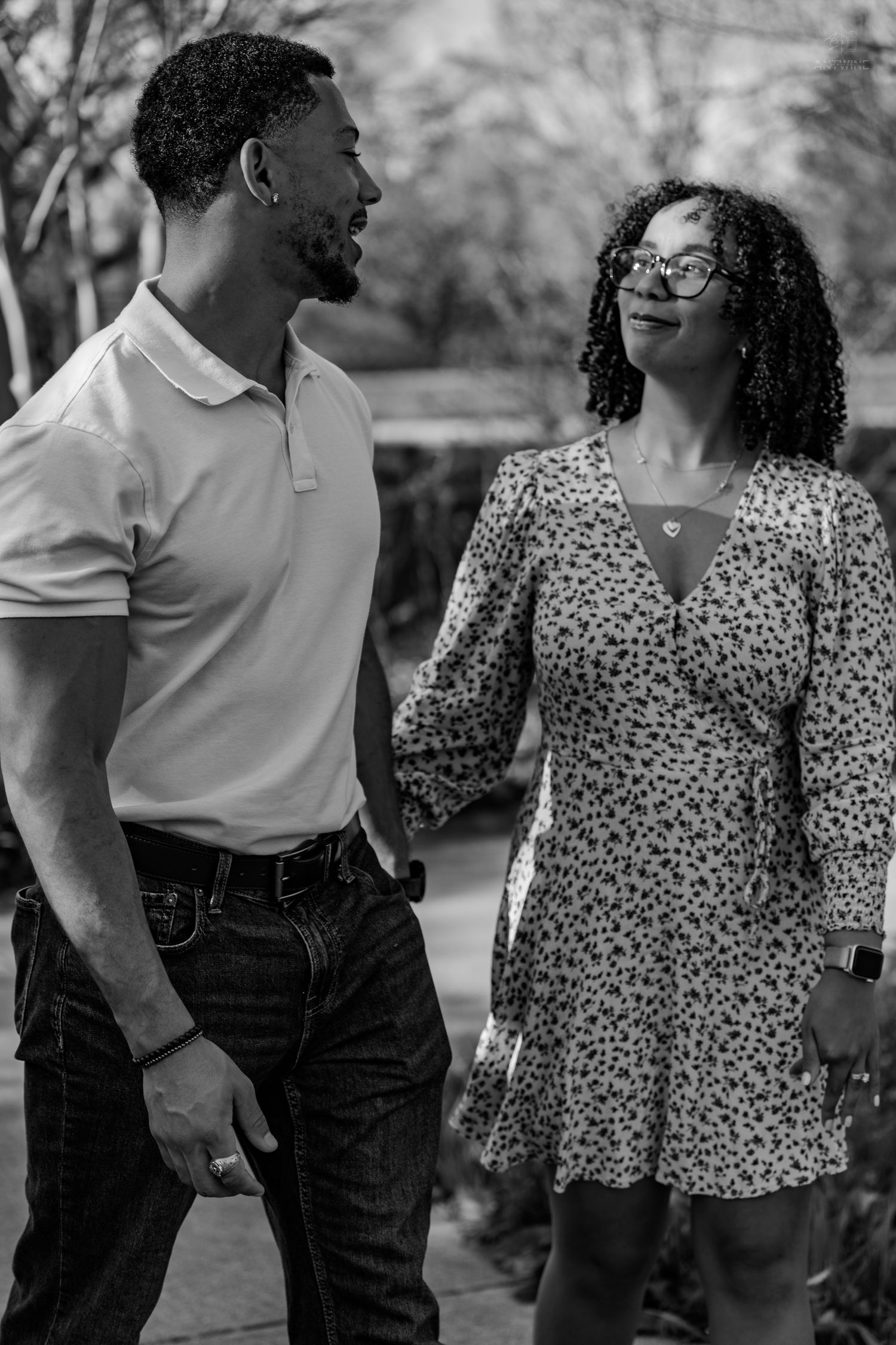 Black and white image of a smiling couple walking and gazing at each other on a sunny park path.
