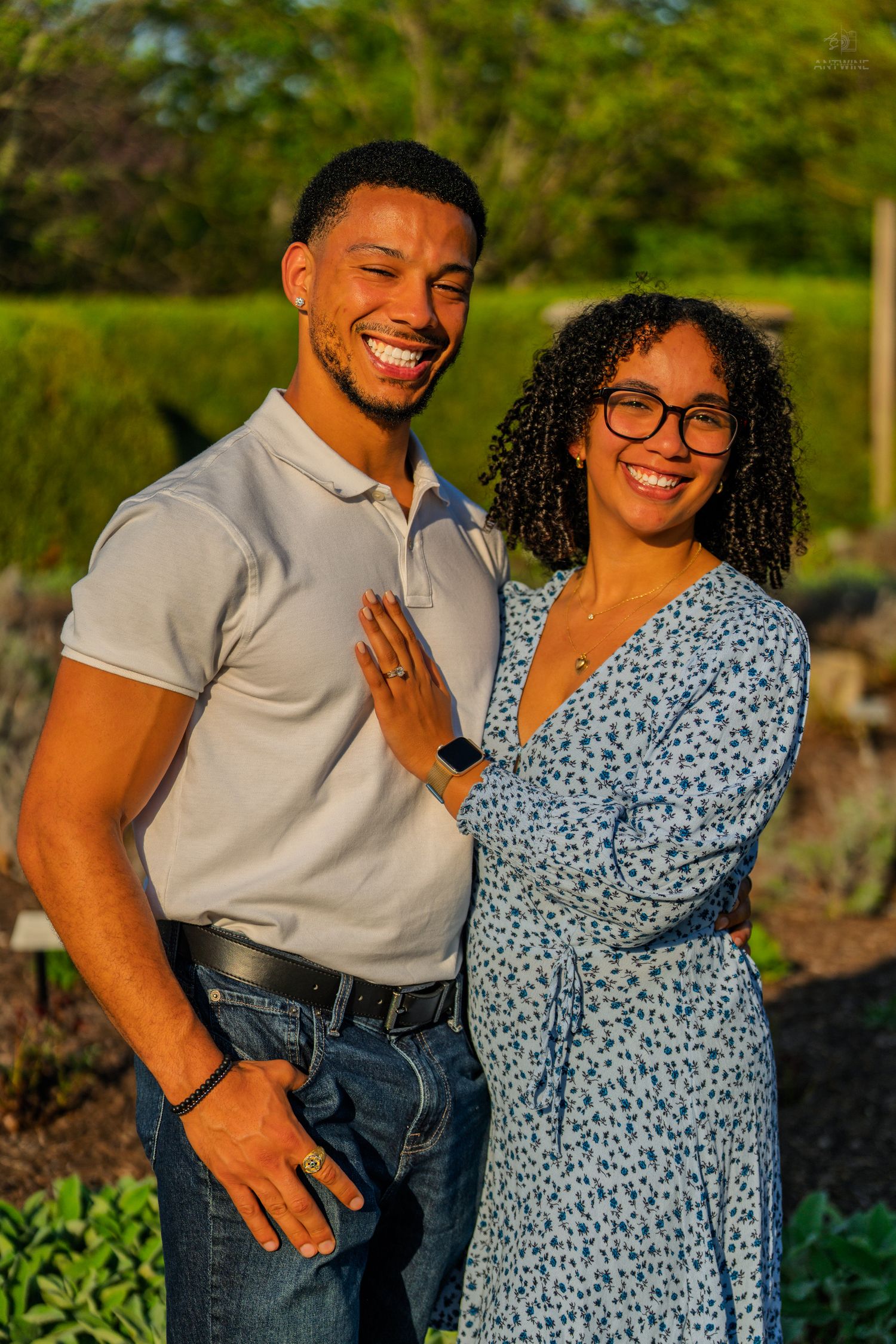 A joyful couple smiles together in a sunlit garden, woman in blue floral dress resting hand on mans chest.