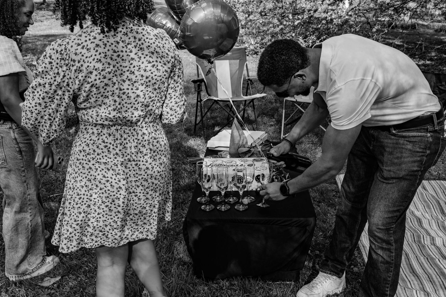 Black and white photo of couple preparing champagne glasses at decorated outdoor picnic table.