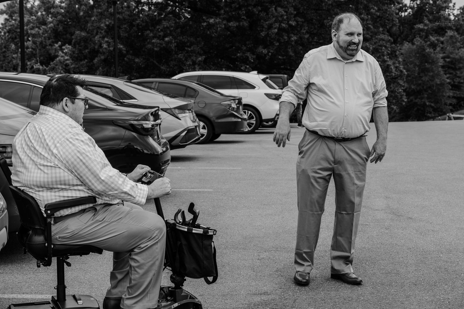 Black and white photo of two men conversing in a parking lot, one seated on a mobility scooter.