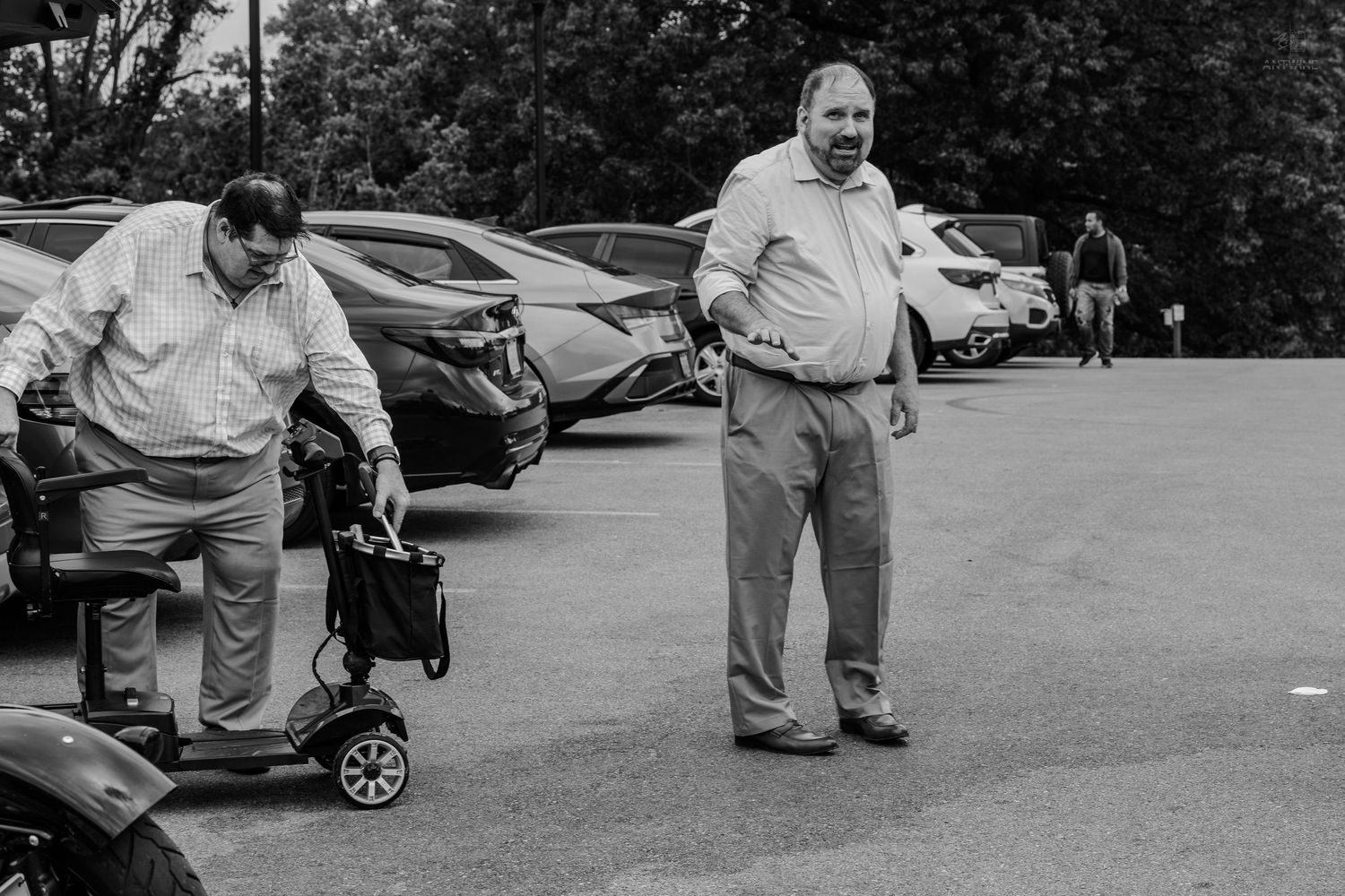 Black and white photo of two men in a parking lot, one handling a mobility scooter.