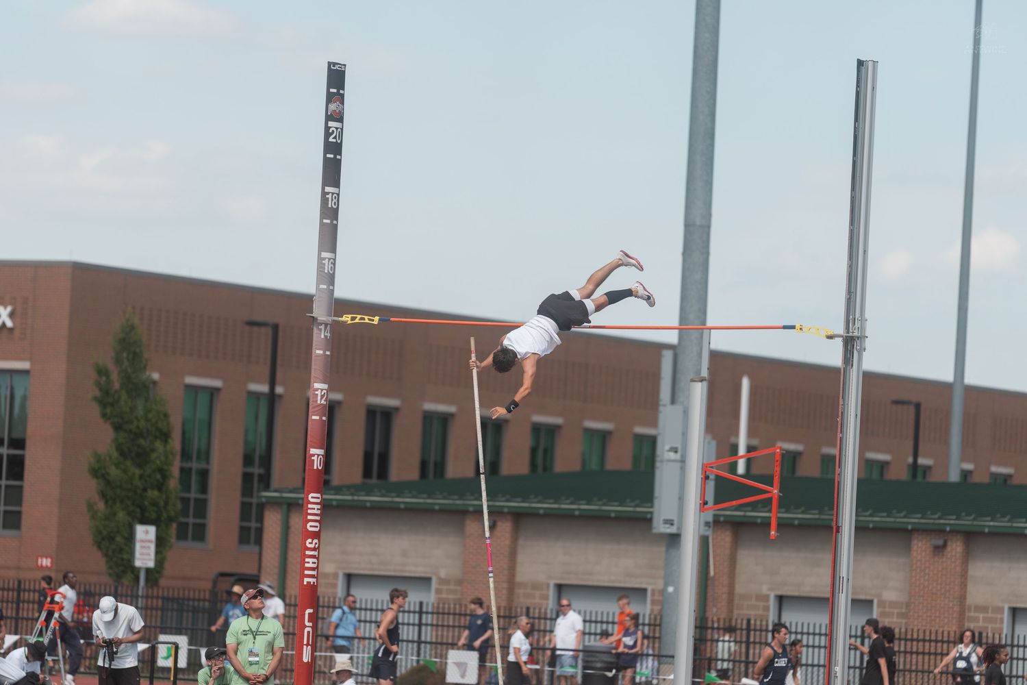 Athlete performs a pole vault jump at an outdoor track and field competition with a red brick building behind.