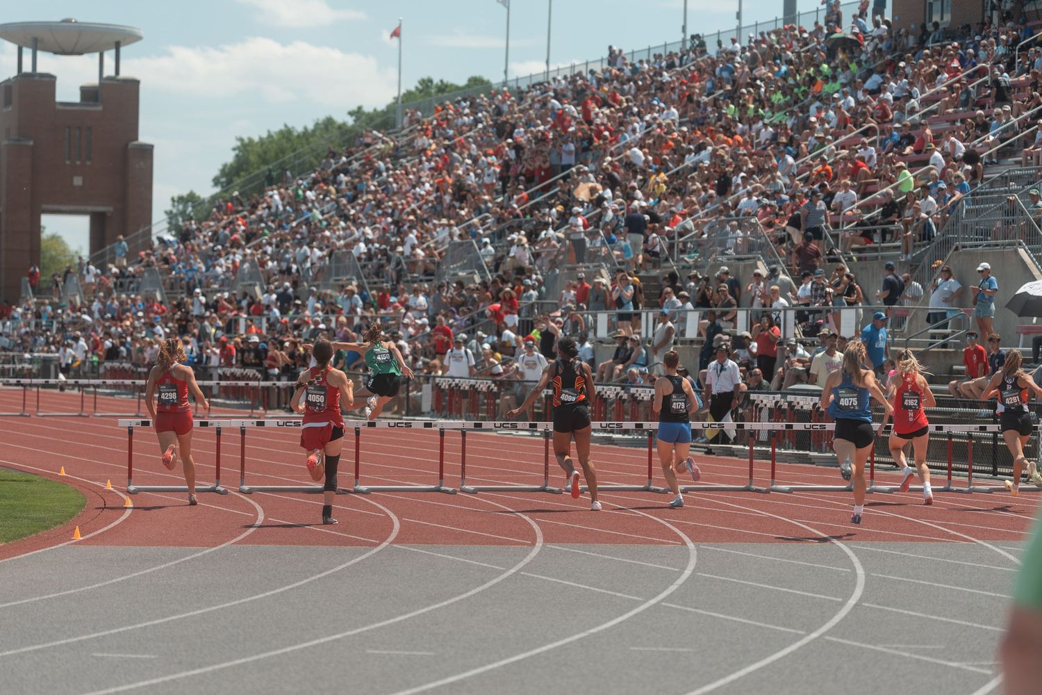 Competitors race over hurdles on a red track at a well-attended high school track and field championship event.