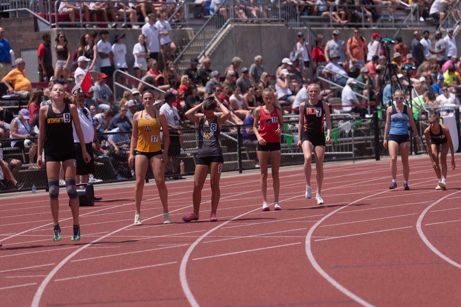 Female track athletes line up at the starting line during a competitive outdoor track and field meet with packed stands.
