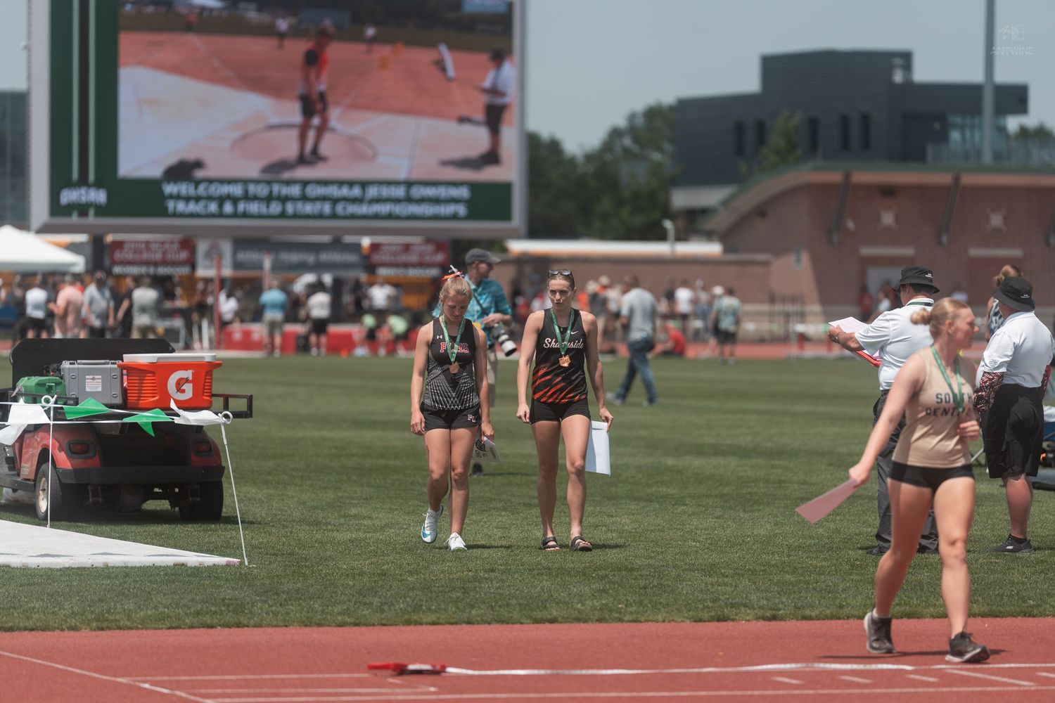 Female athletes carry javelins across the infield at the OHSAA Track and Field State Championships on a bright sunny day.