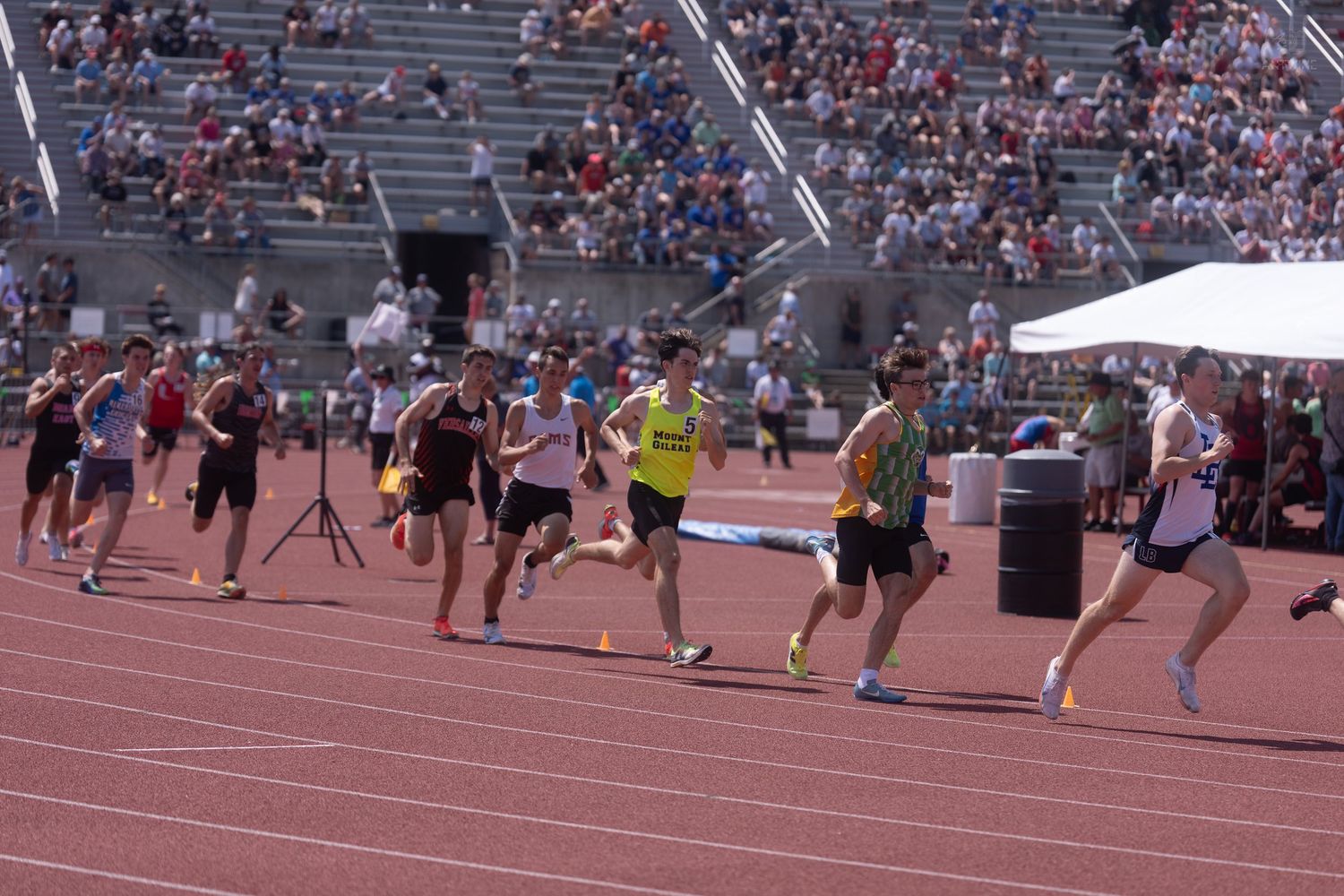 Male athletes sprint down the track during a competitive outdoor track and field meet on a sunny day.