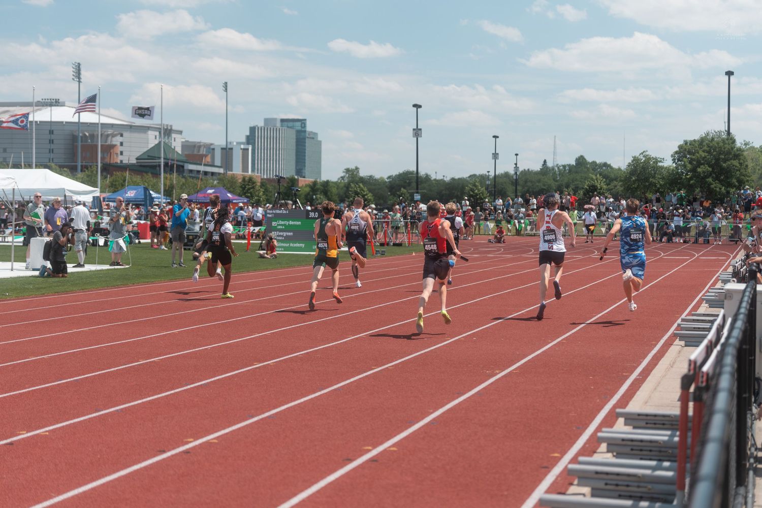 Multiple athletes race down a red outdoor track during a sunny day at a competitive track and field meet.