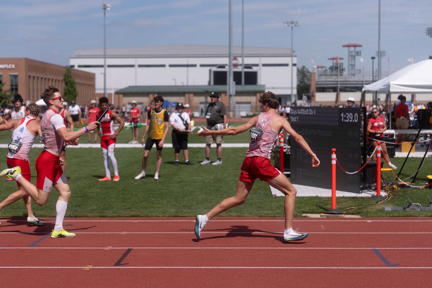Two relay runners complete a baton exchange on a red track at an outdoor high school track and field meet.