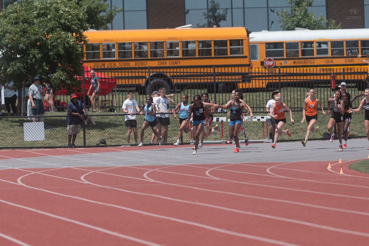 Female runners sprinting past yellow school bus at outdoor high school track and field competition.