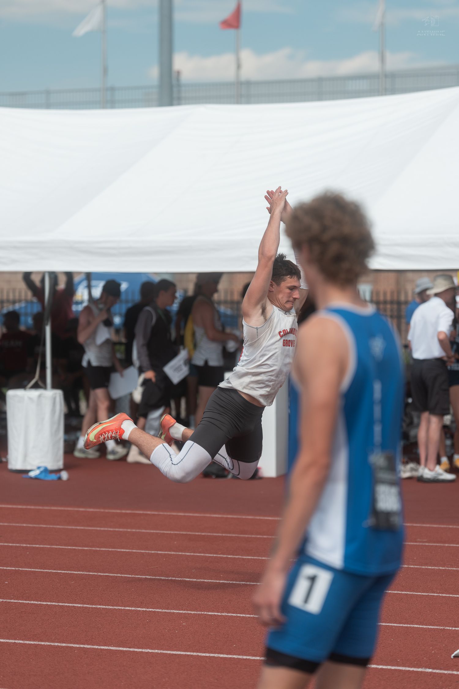 Athlete executes a long jump with arms raised at an outdoor track and field competition on a sunny day.