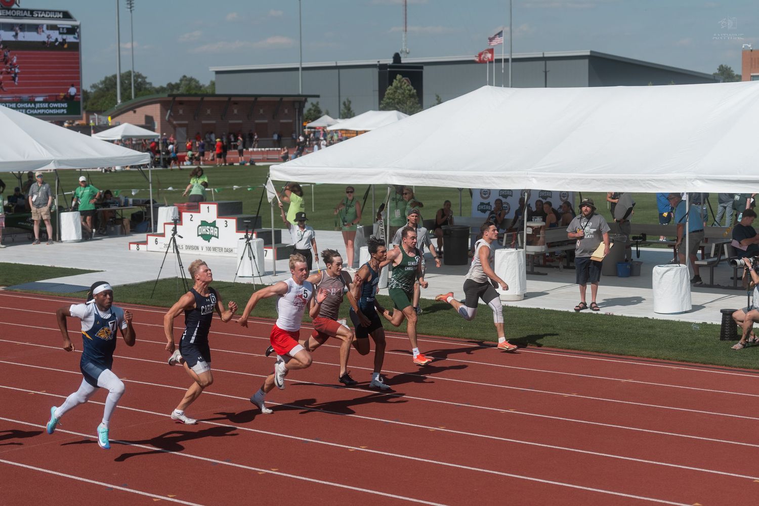 Track athletes in colorful uniforms race on a red track during an outdoor athletic competition on a sunny day.