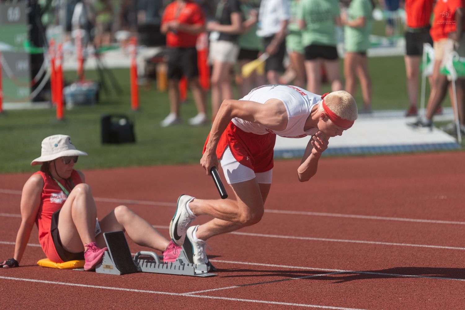 Senior male athlete in red and white uniform launches from starting blocks at outdoor track competition.