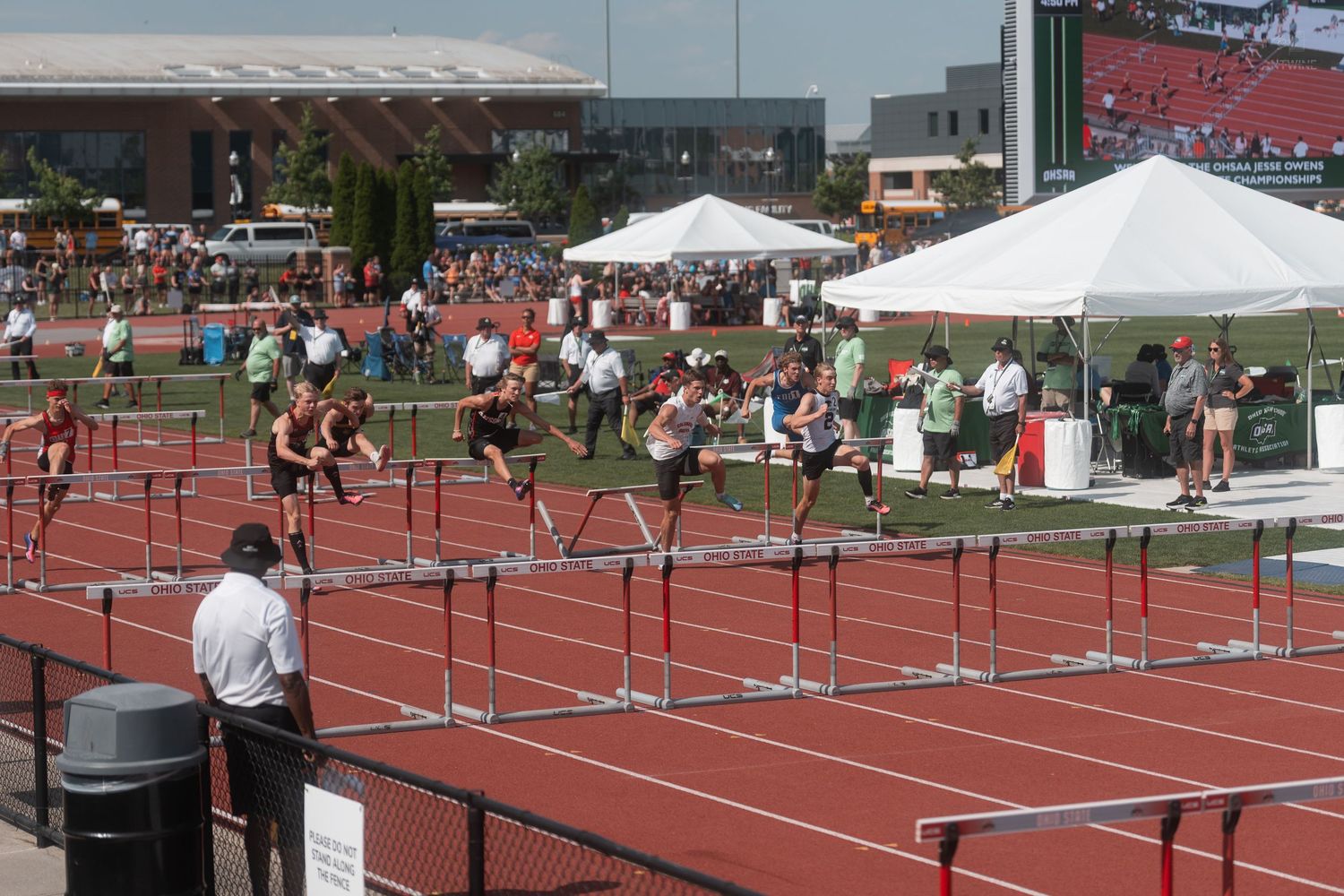 Athletes competing in hurdles race on red track at outdoor high school championship with white tents and crowd.