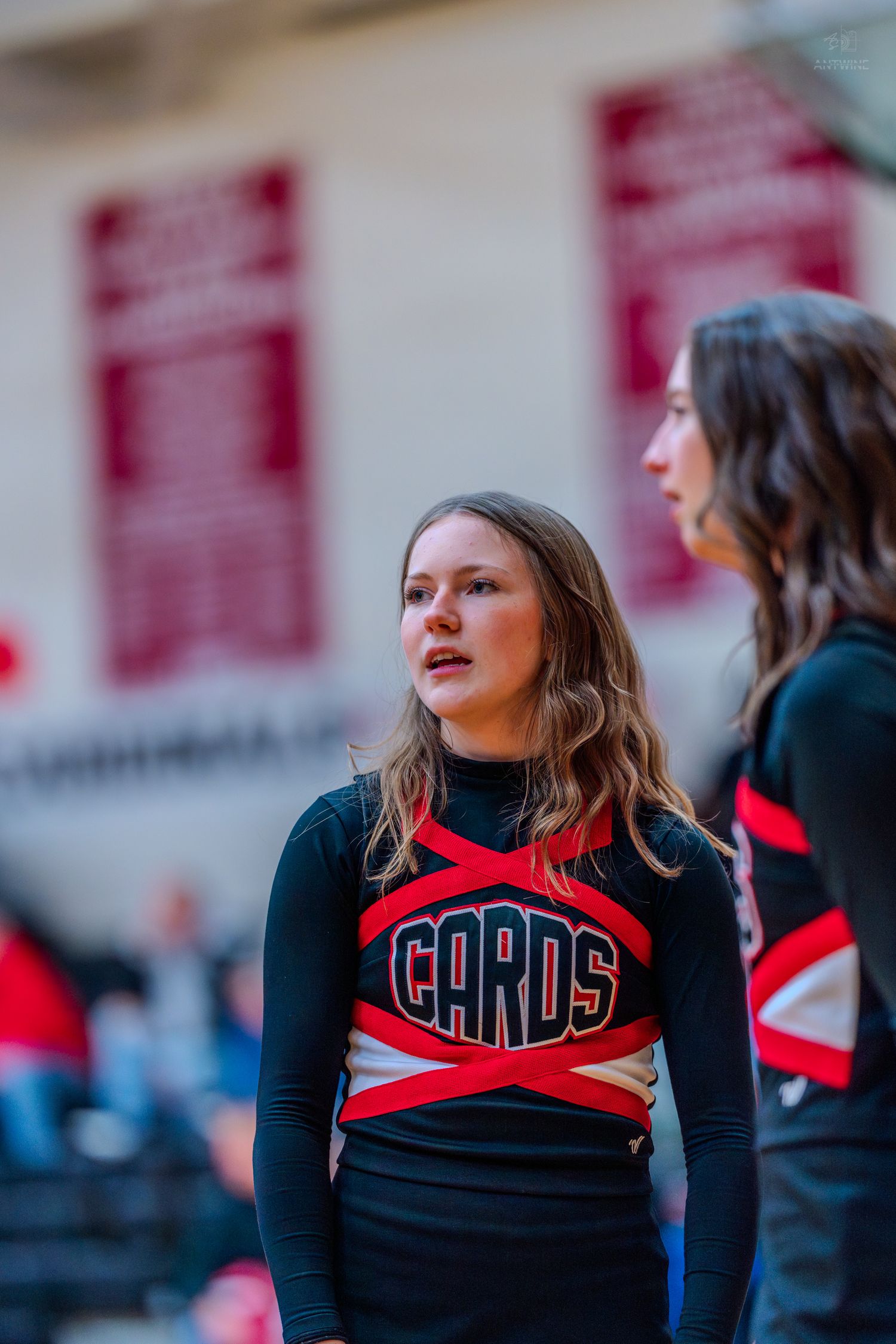 High school cheerleader in black and red CARDS uniform stands attentively beside teammate in gymnasium.