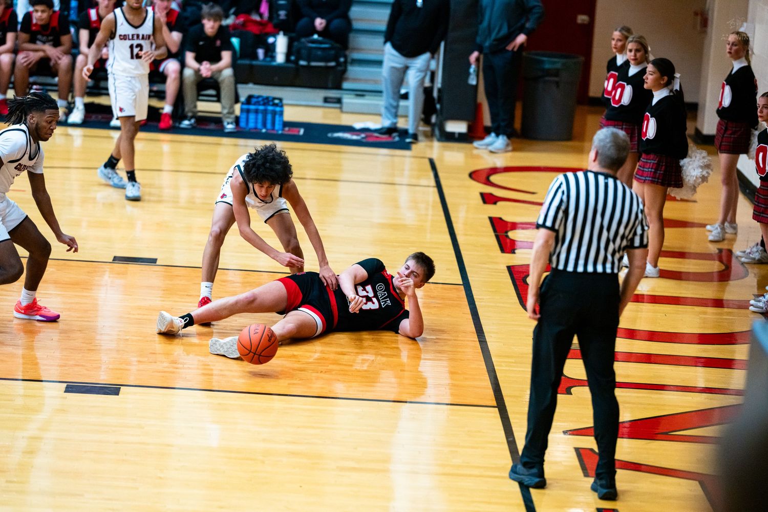 Basketball player lies on court floor reaching for loose ball as referee watches closely.