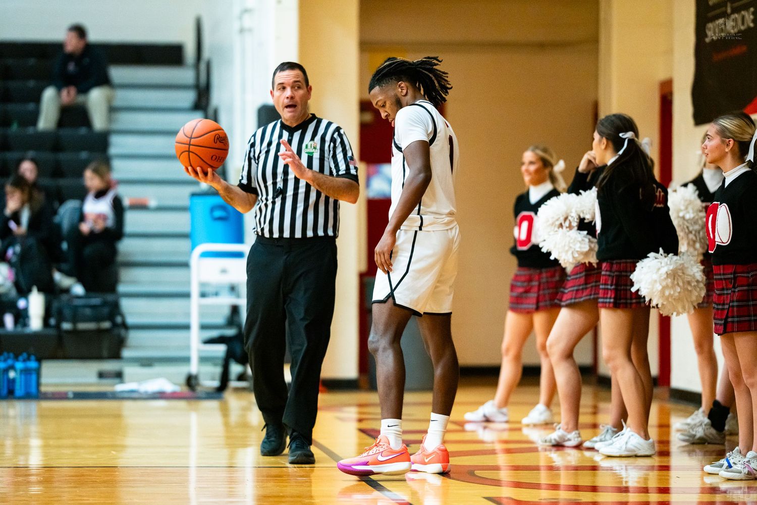 Referee hands basketball to tall player near cheerleaders during high school basketball game.