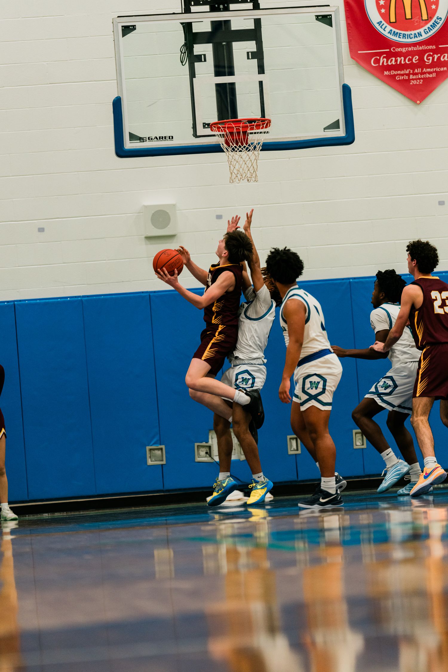 A Turpin player in maroon drives to the basket for a layup against defenders in white during a gym game.