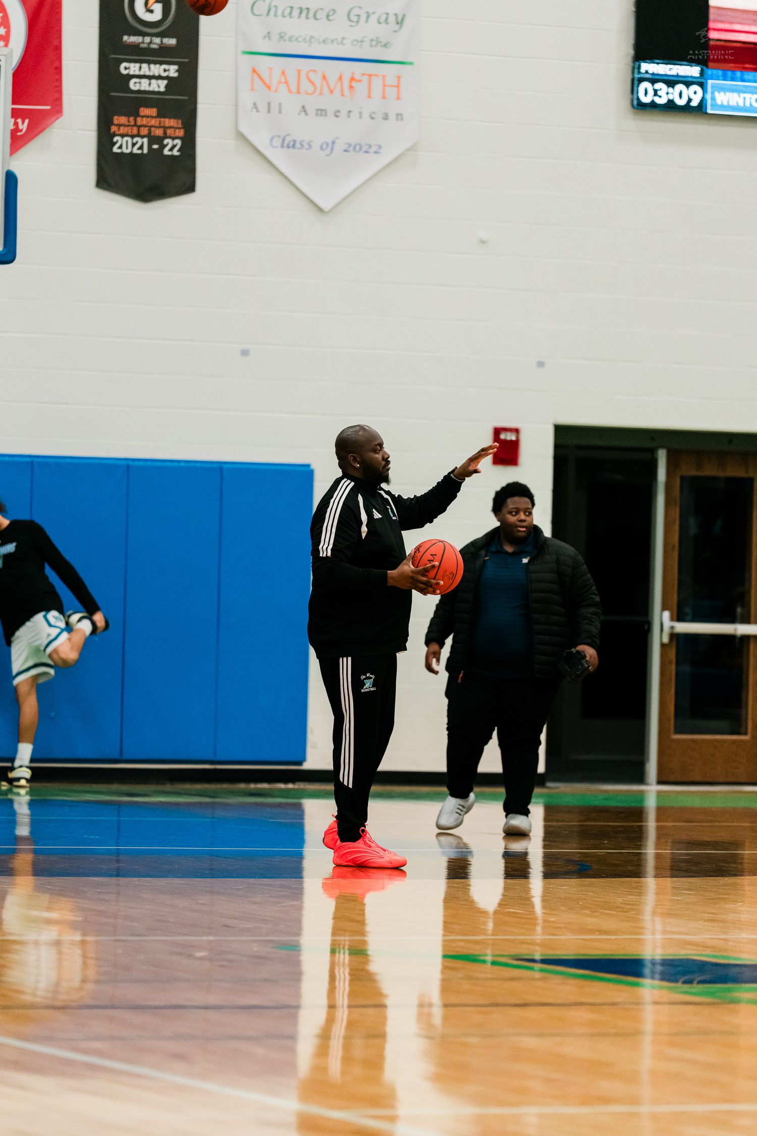 A basketball coach holds a ball and points during a practice session in a school gymnasium.