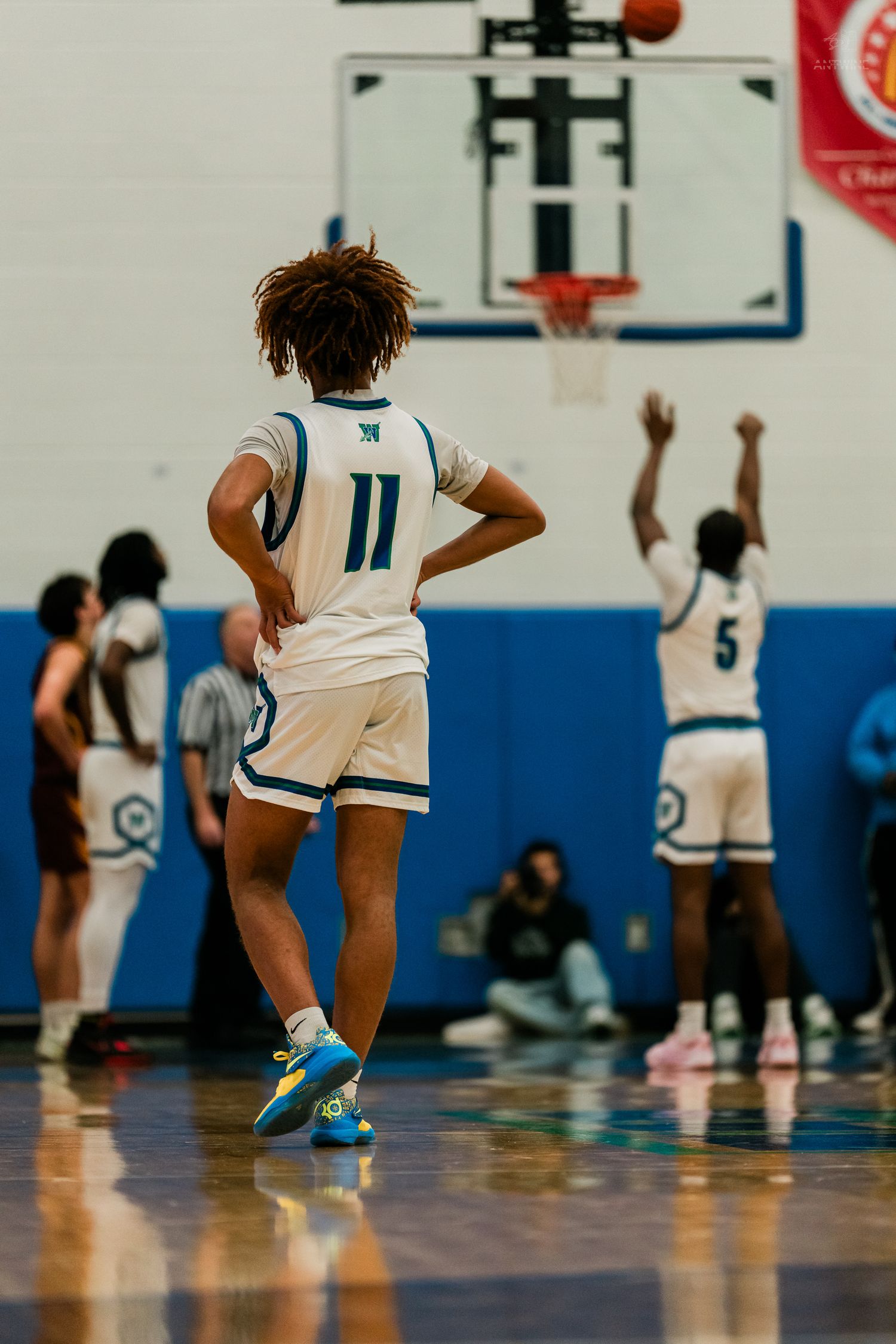 Basketball player wearing number 11 white Warriors jersey watches teammate attempt a free throw shot.