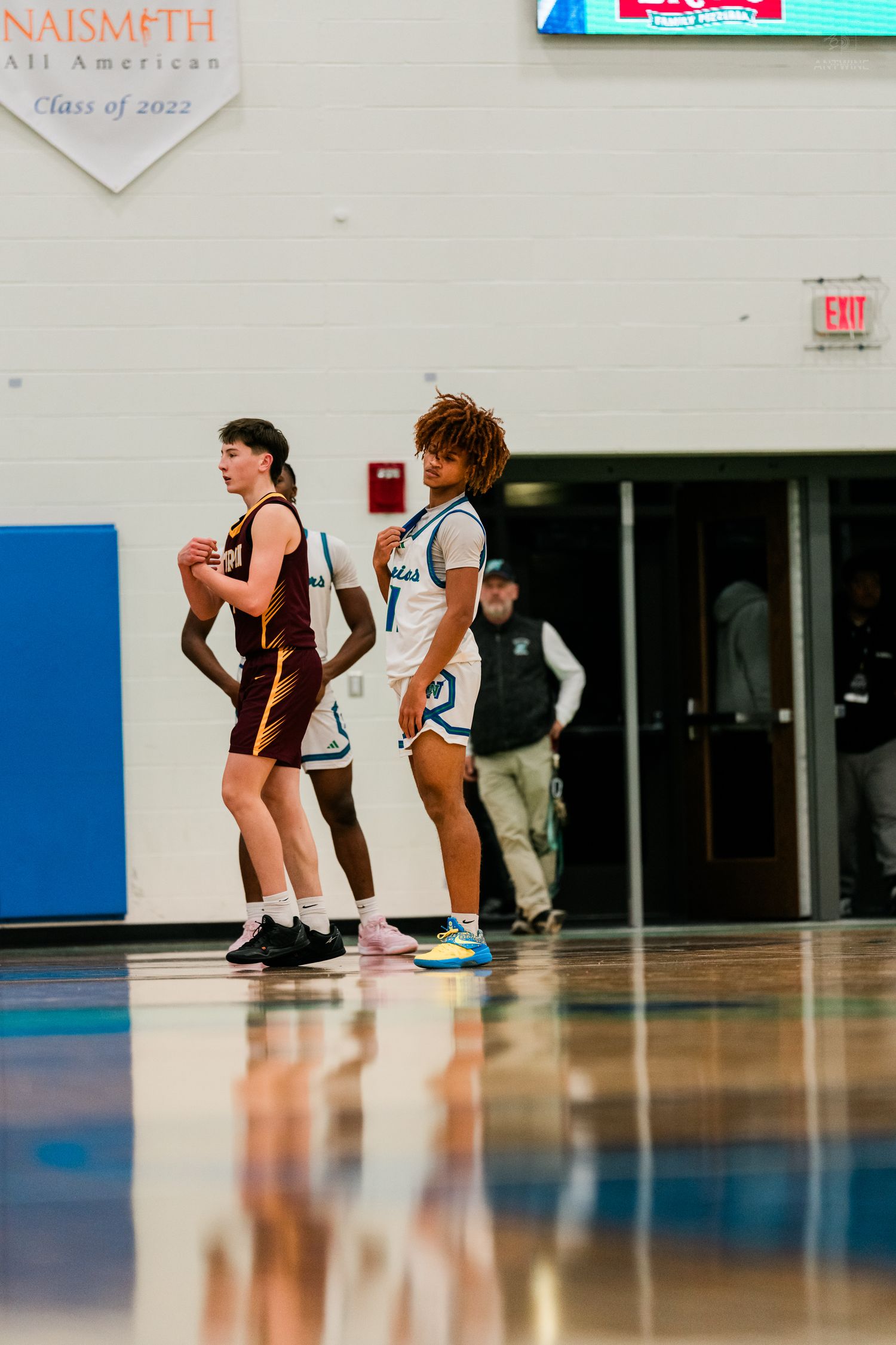 Two female basketball players stand on court during a high school game in a blue and white gymnasium.