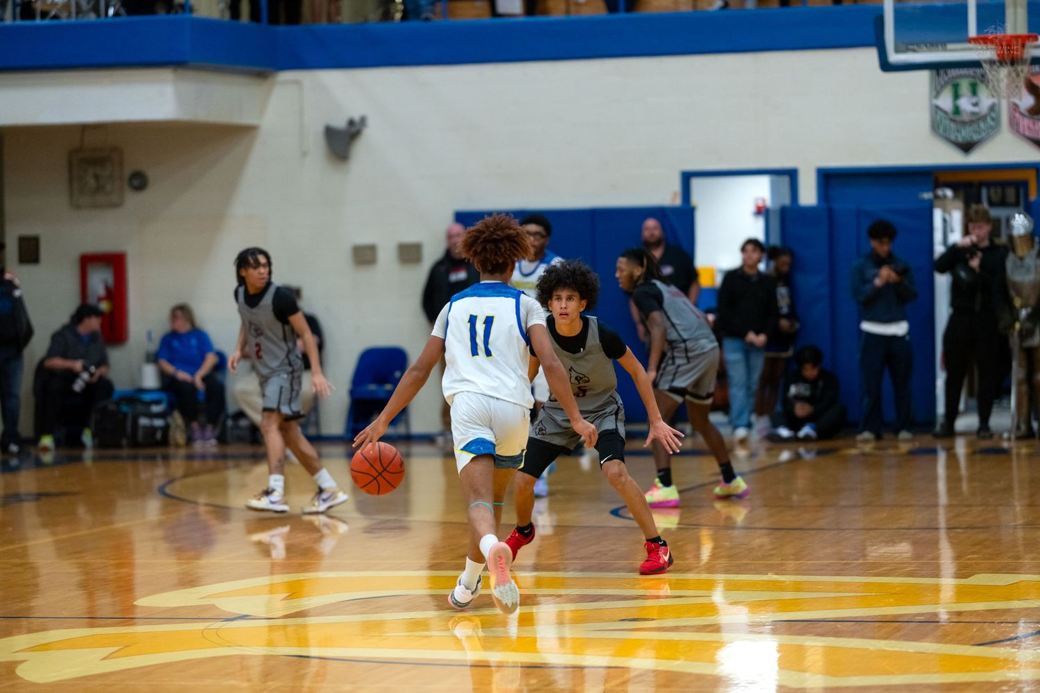 High school basketball player in white number 11 jersey dribbles past a defender on a shiny gymnasium court.