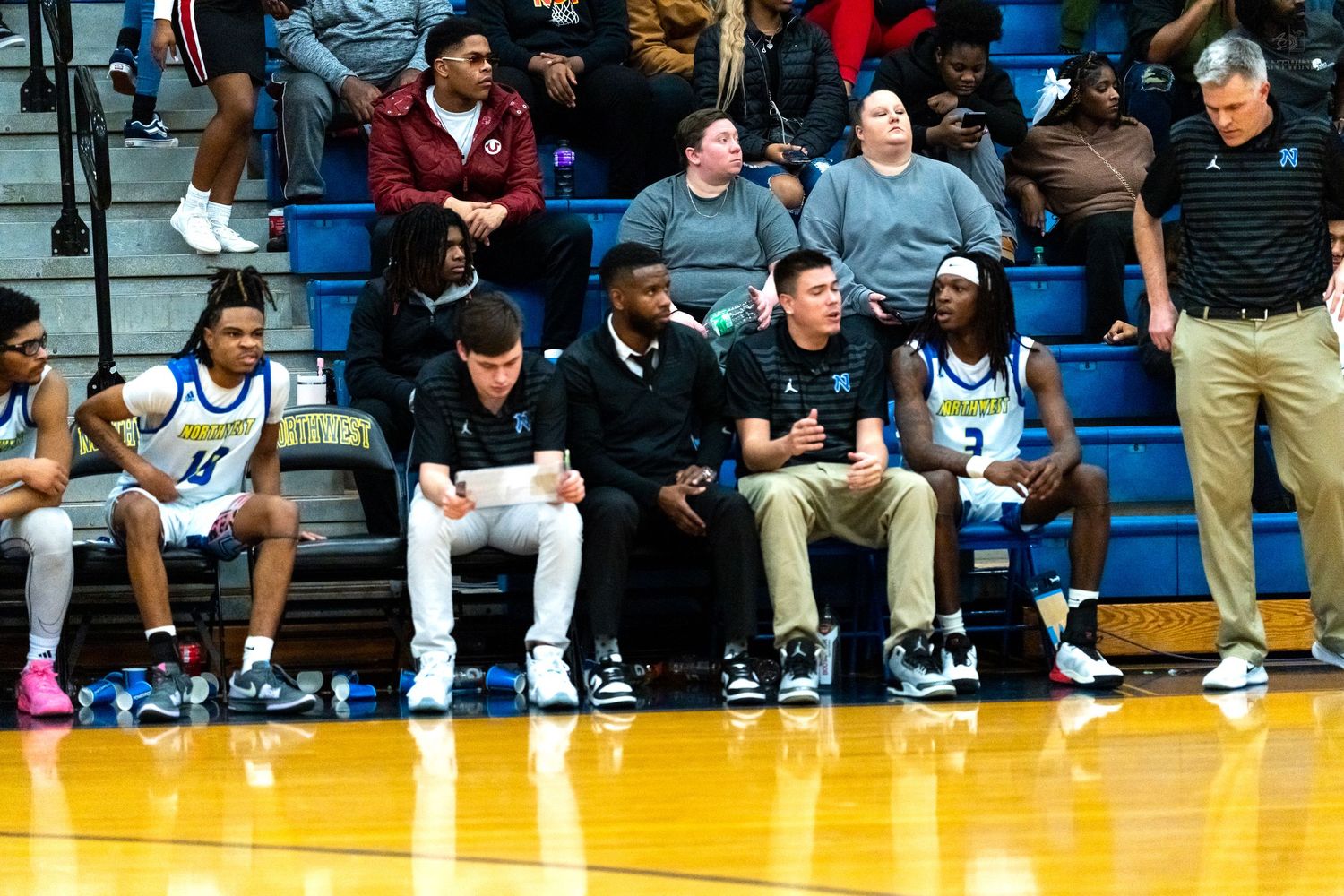 High school basketball team sits on the bench with coaches during a game in a packed gymnasium.