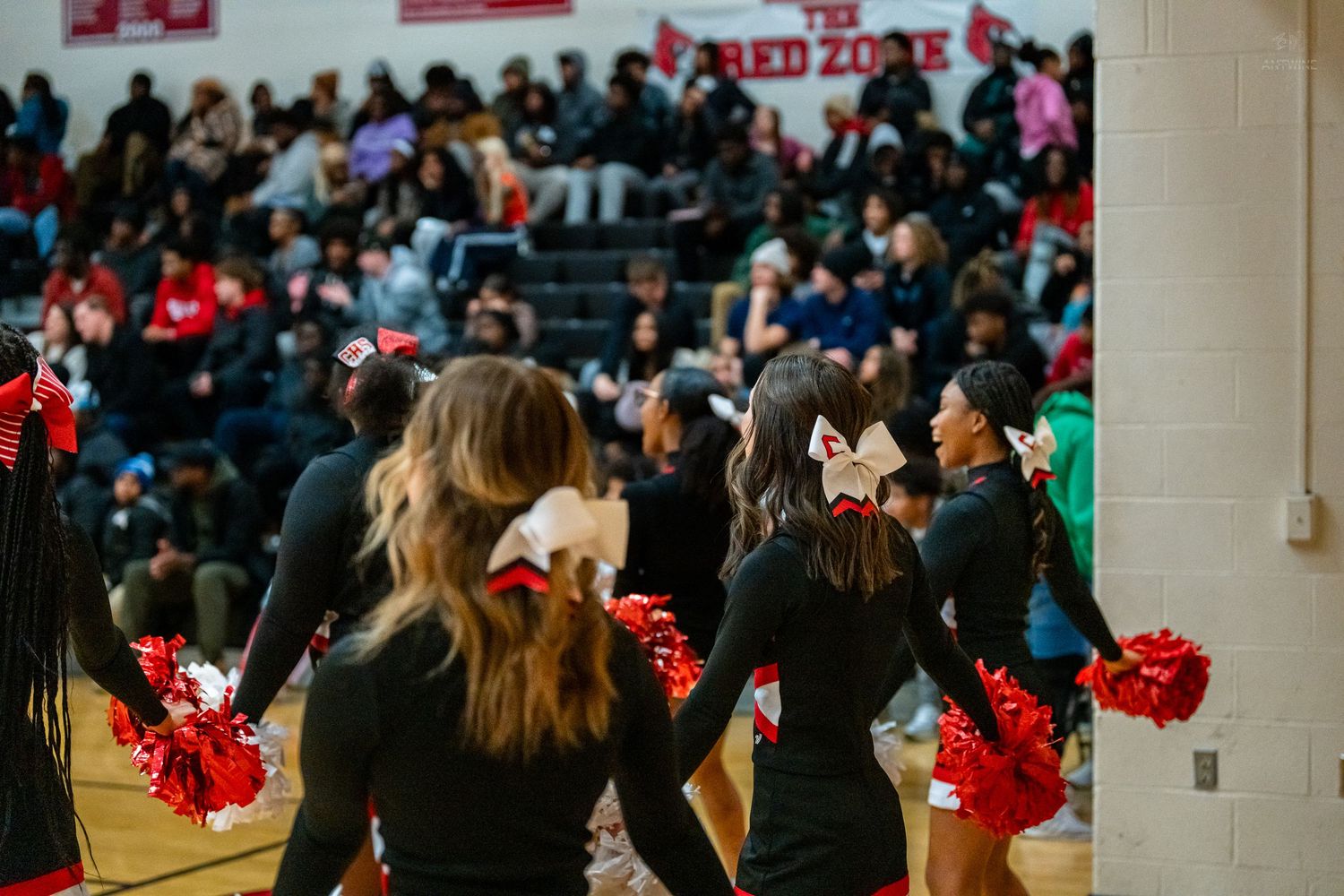 Cheerleaders in black uniforms with red pom-poms perform for a packed crowd at a high school basketball game.