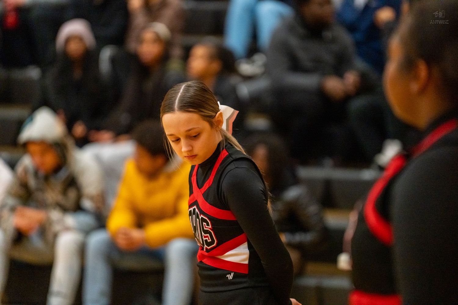 A young cheerleader in a black and red uniform looks down during a high school basketball game.
