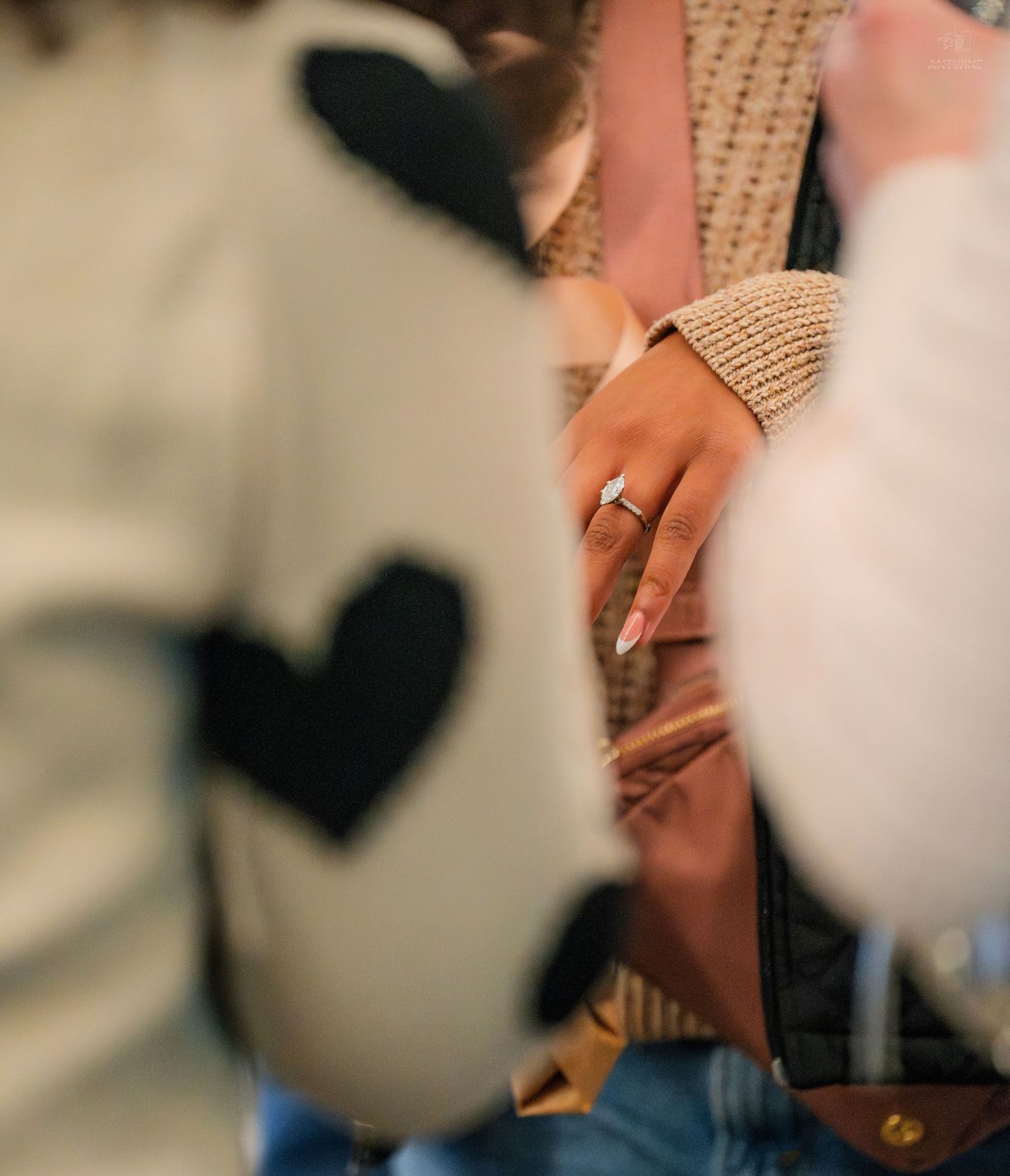 Close-up of a woman's hand displaying a diamond engagement ring at a social gathering.