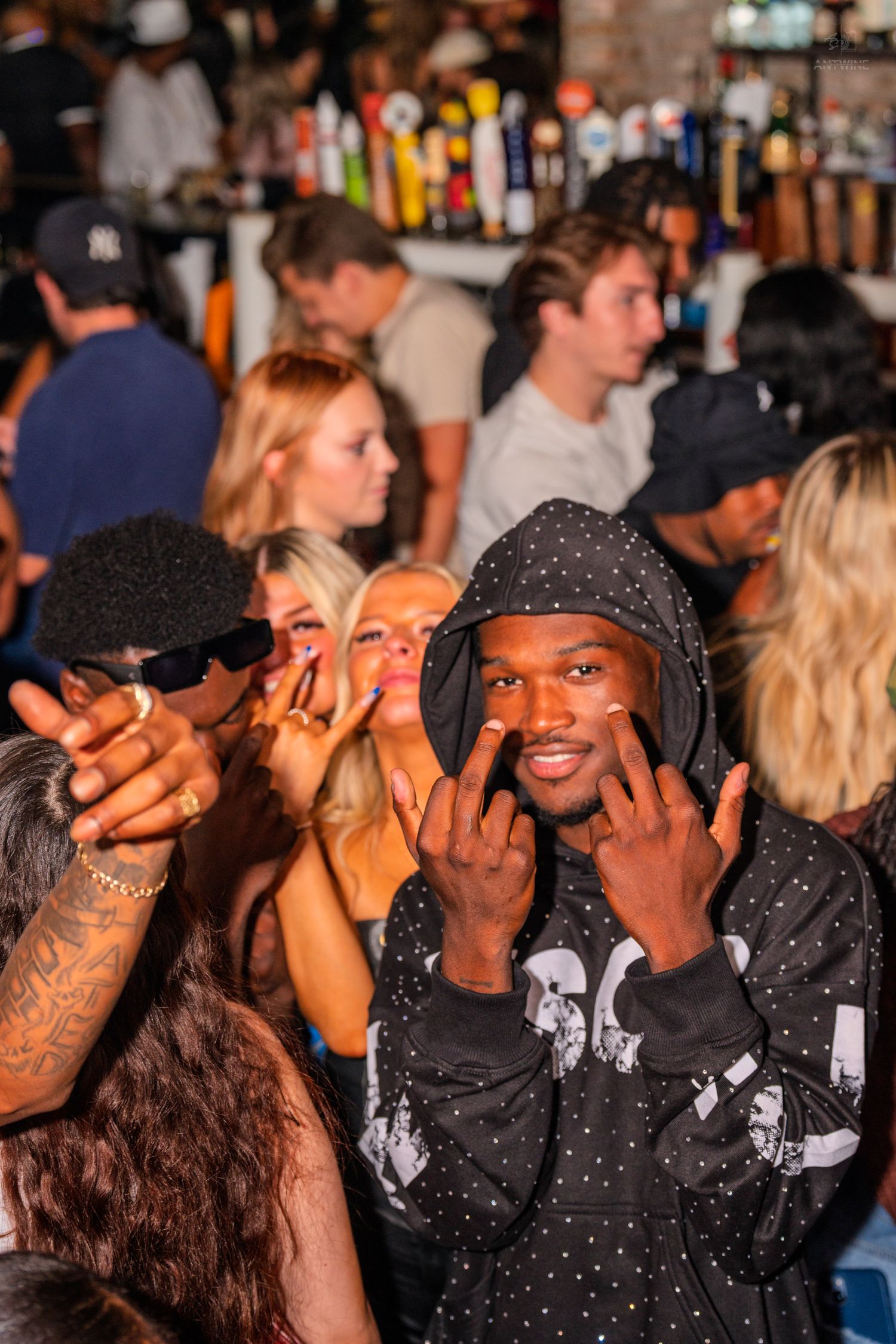 Young man in hooded sweatshirt making hand gestures at a busy bar with beer taps.
