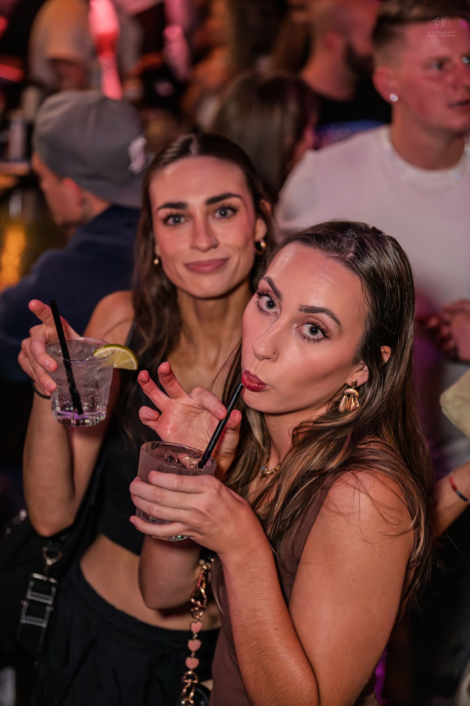 Two women posing with cocktails and smiling at a crowded nightclub party.