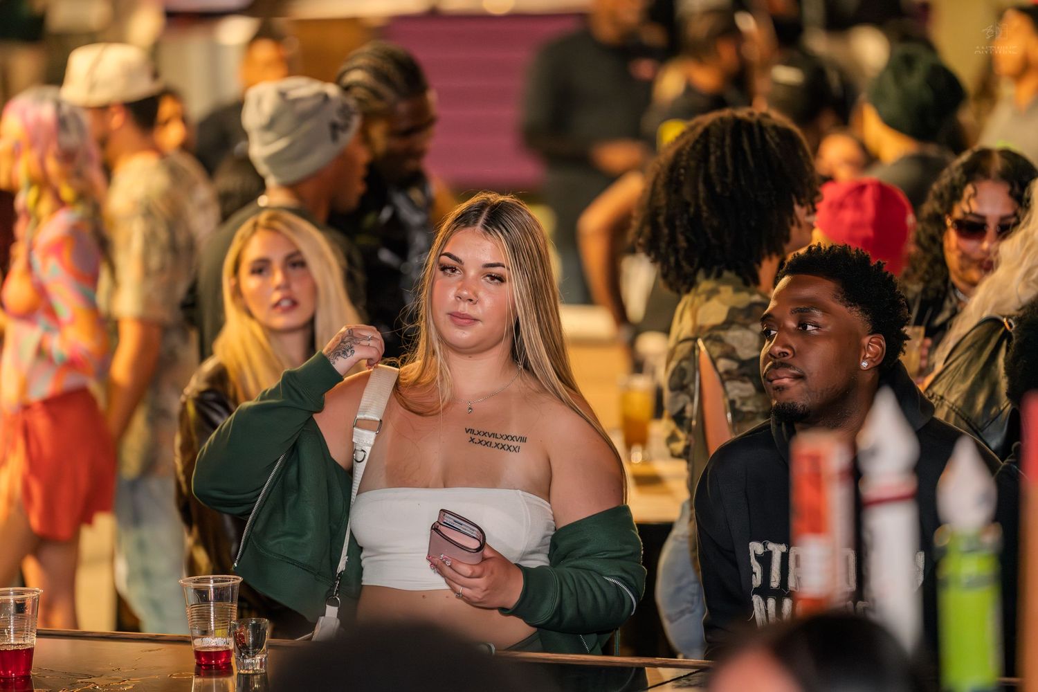Young woman in white top holding a phone sits at a busy bar surrounded by a diverse crowd.