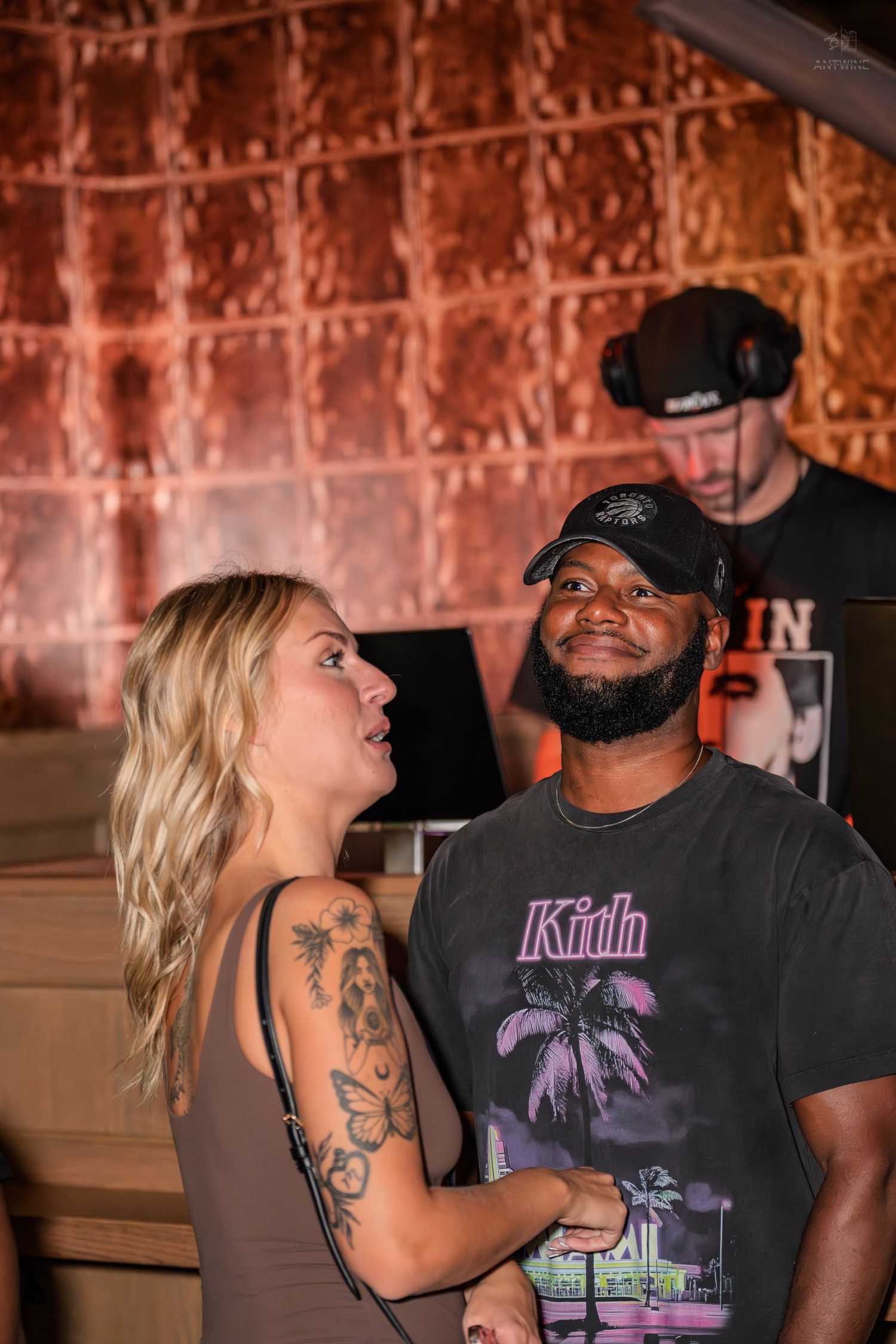 A smiling couple poses together at a nightclub with a DJ visible in the background.