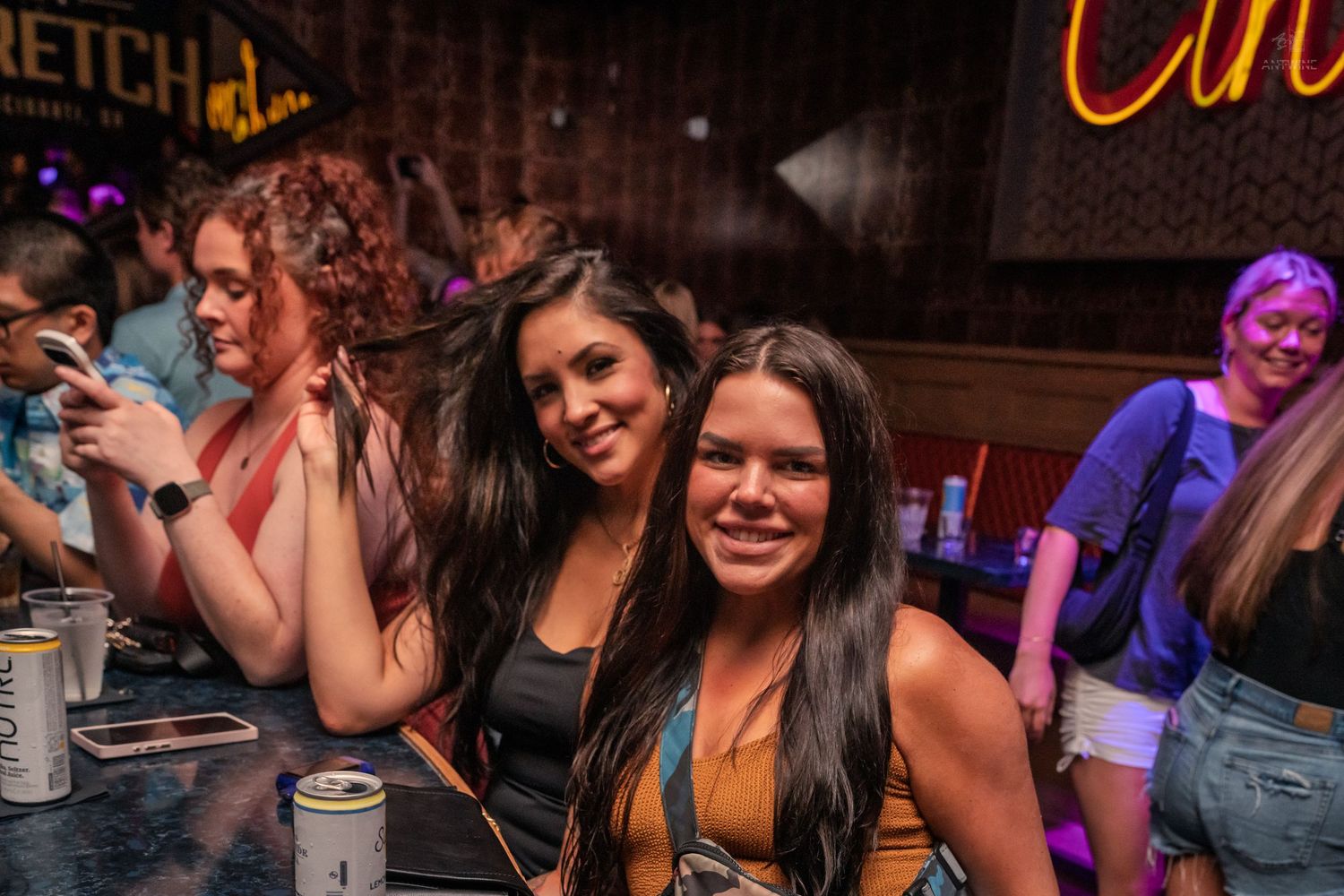 Two smiling women pose together at a lively bar with neon signs and drinks on the table.