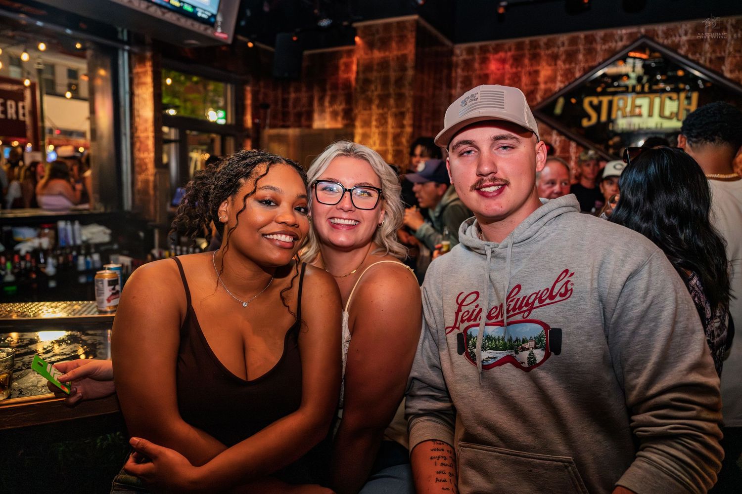 Three friends posing and smiling together at a crowded bar beneath a Stretch sign.