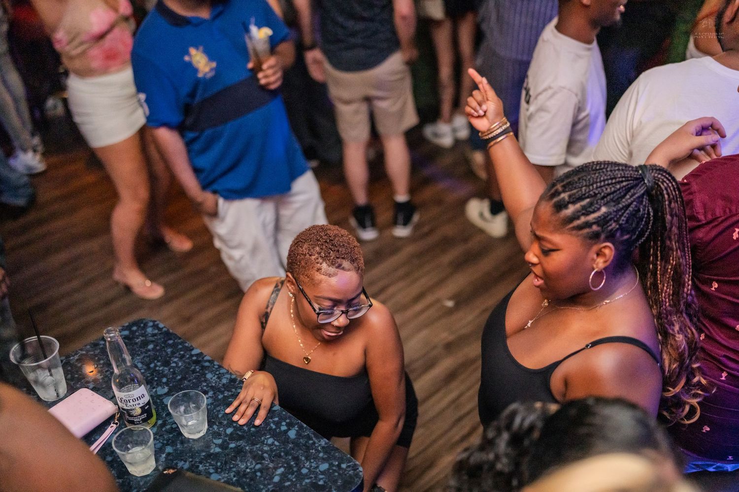 Two women dancing and celebrating at a nightclub bar table with drinks during a party event.