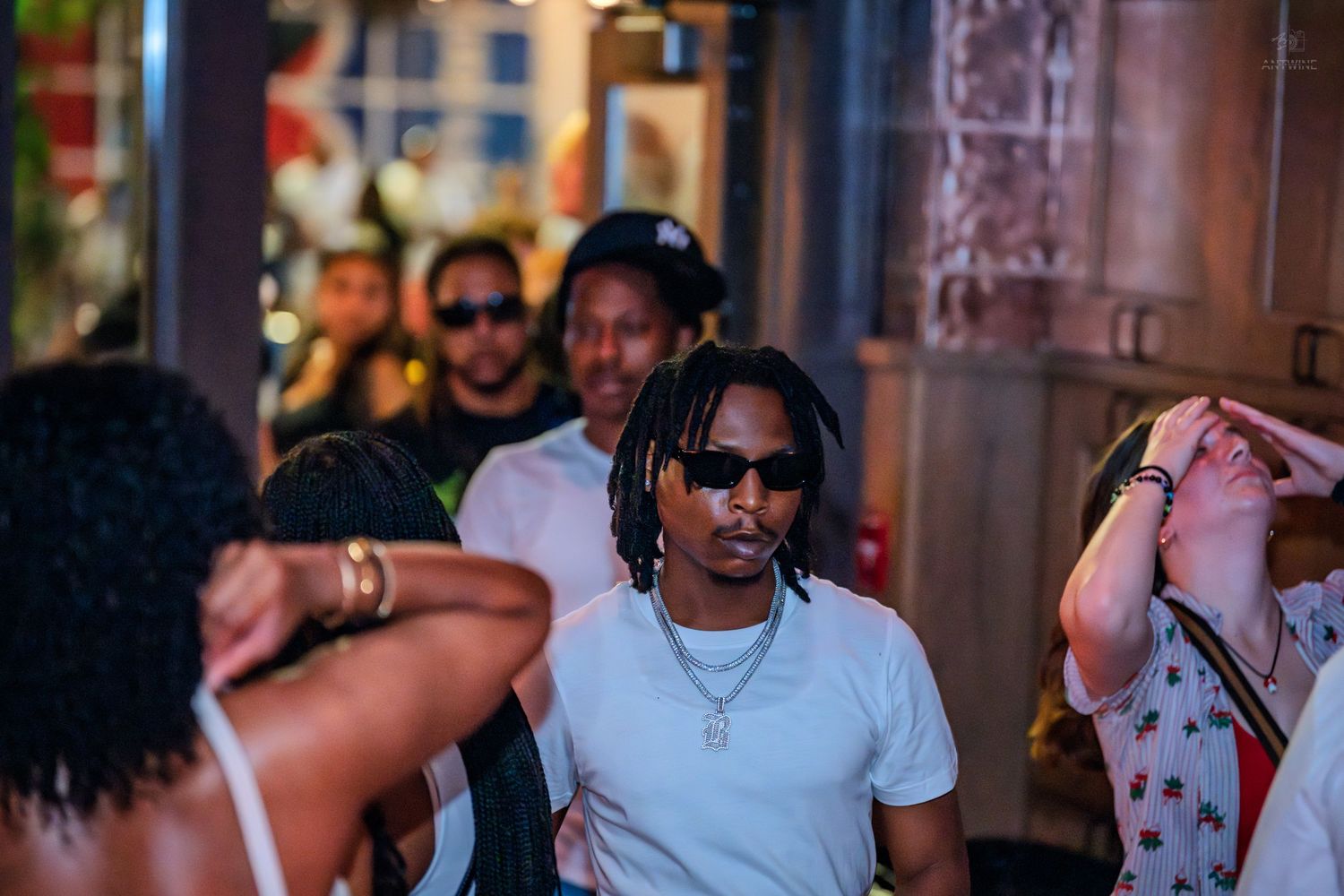 Man in white shirt with chain necklace and sunglasses at crowded nightclub.