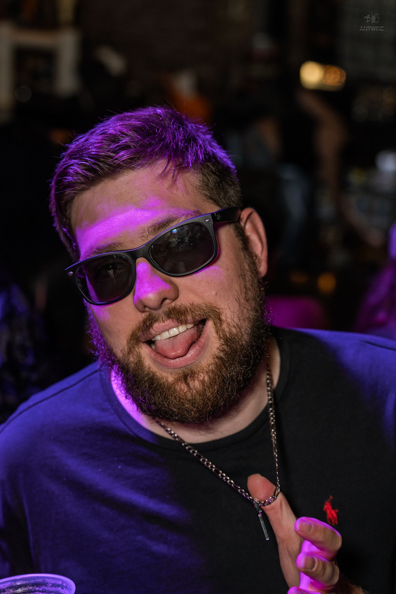 Bearded man wearing sunglasses and black shirt smiling under purple lights at a nightclub.