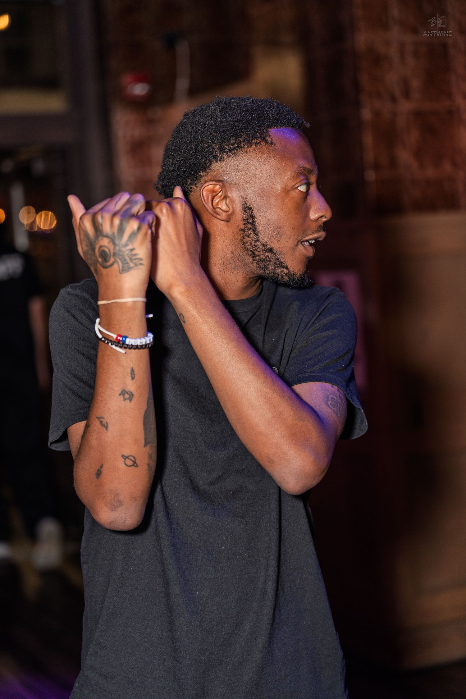 Tattooed man in a black t-shirt with bracelets posing with hands raised at a nightclub.