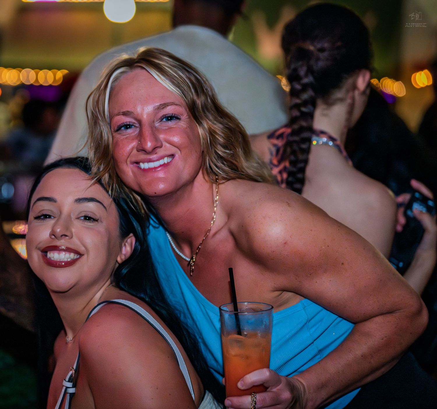 Two women smiling and posing together at a bar, one holding a tropical cocktail drink.