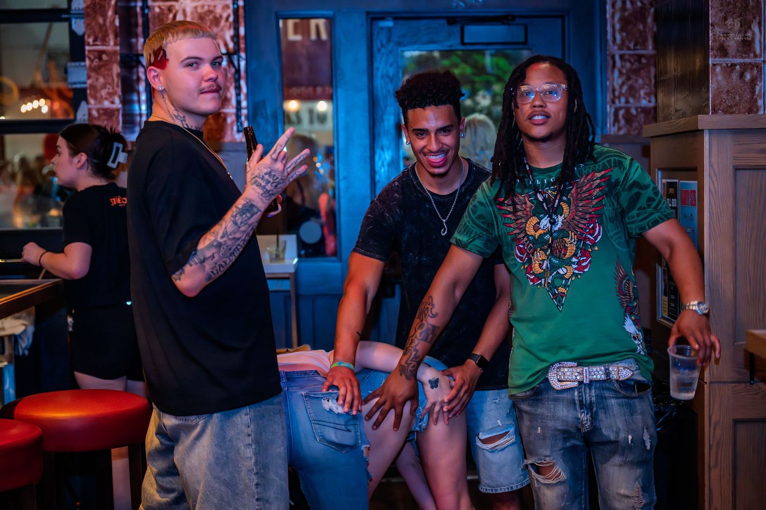 Three men smiling and posing together in a colorfully lit bar or nightclub setting.